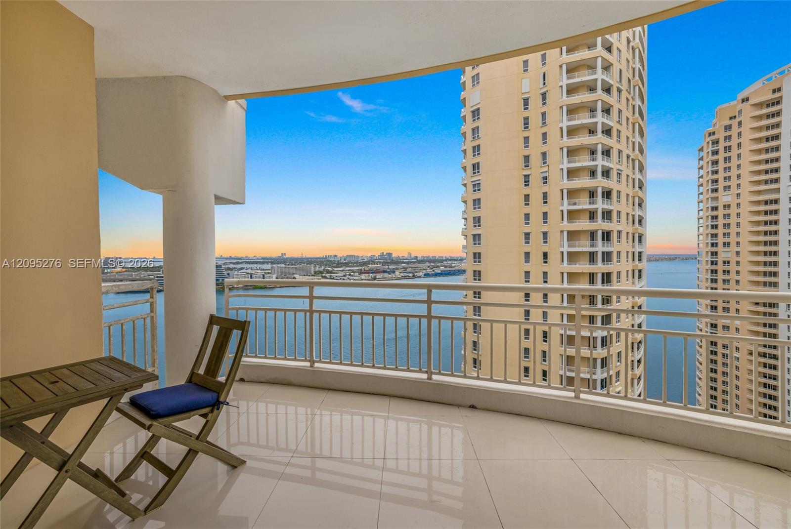 High-rise balcony with chairs overlooking water, neighboring towers, and a distant city skyline at sunset.