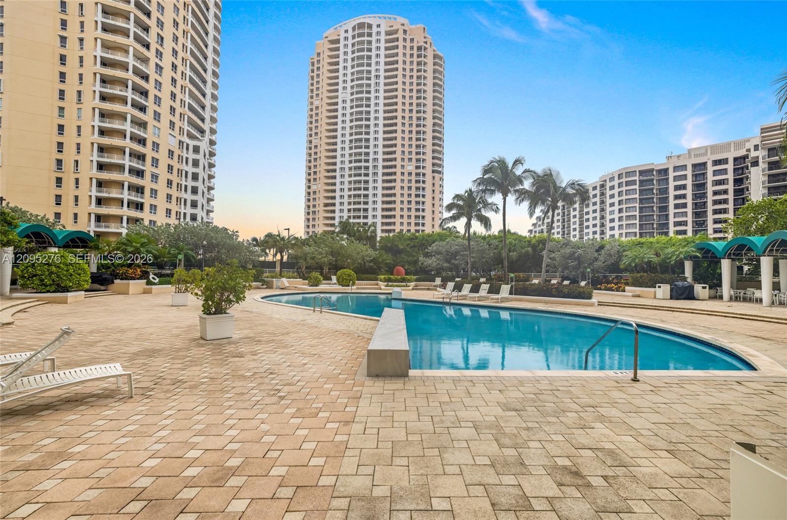 Condominium pool beside a broad paved deck, with palm trees and high-rise buildings.