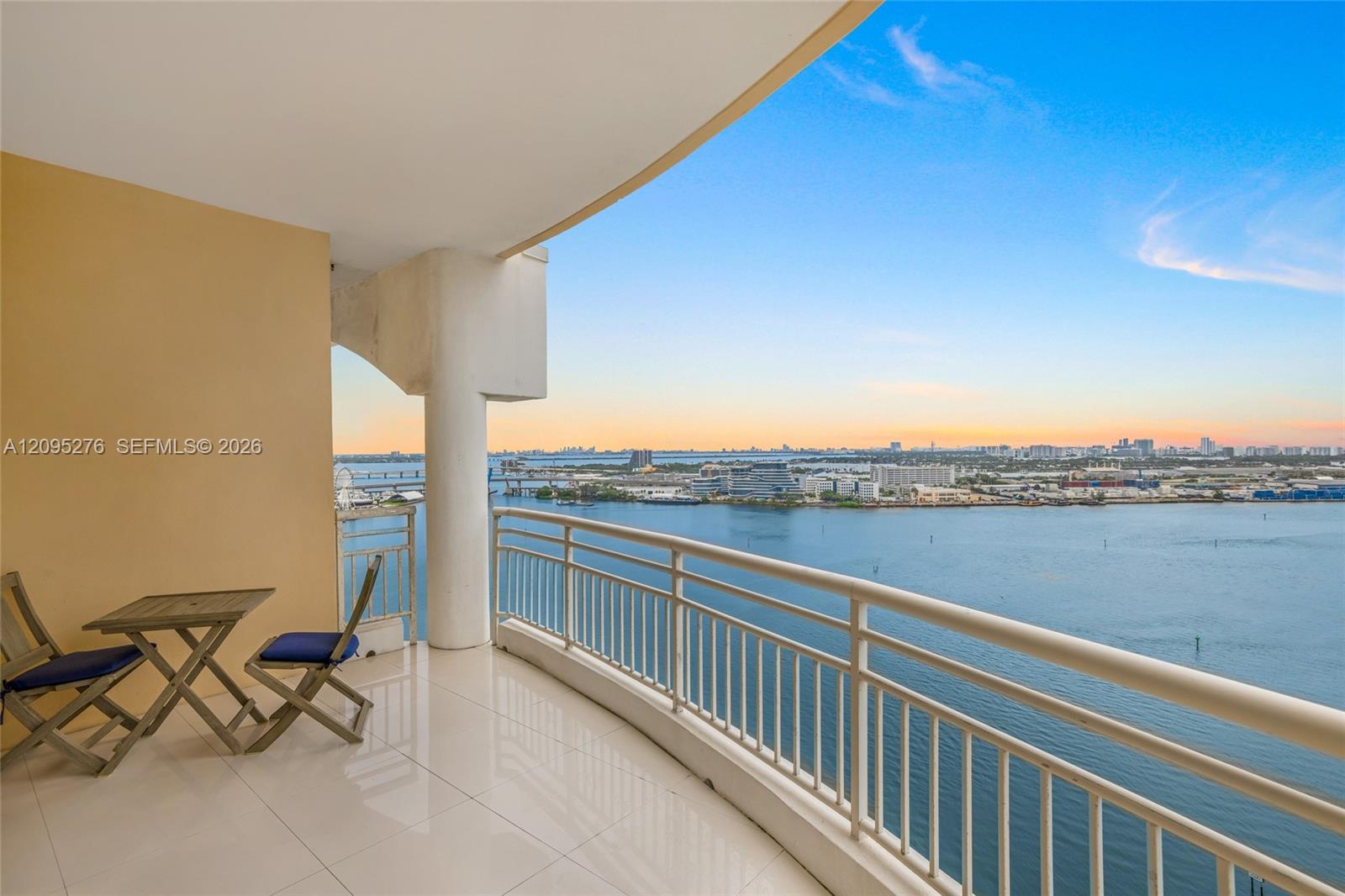 Furnished balcony overlooking a bay, bridges, and a distant skyline at sunset.