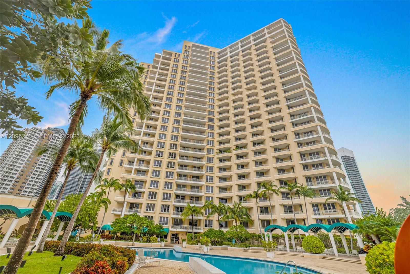 Beige high-rise condominium with balconies, framed by palm trees and a landscaped swimming pool.