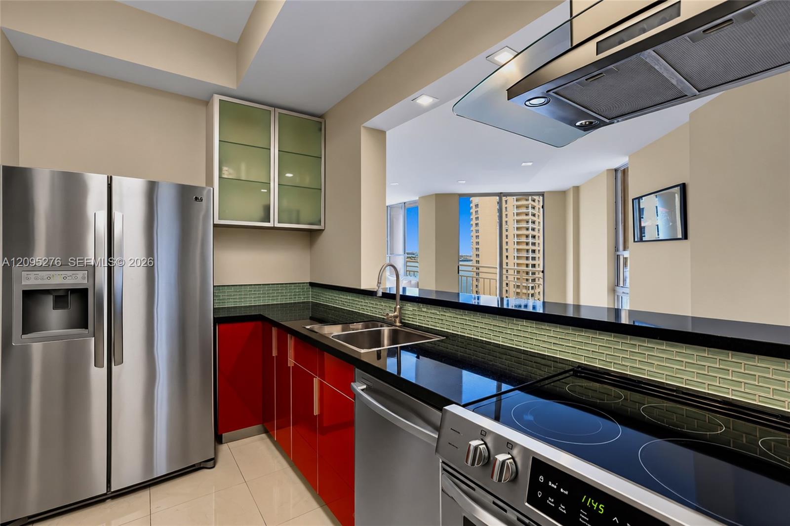 Condo kitchen with red cabinets, green tile backsplash, stainless appliances, and a glimpse of water through the windows.