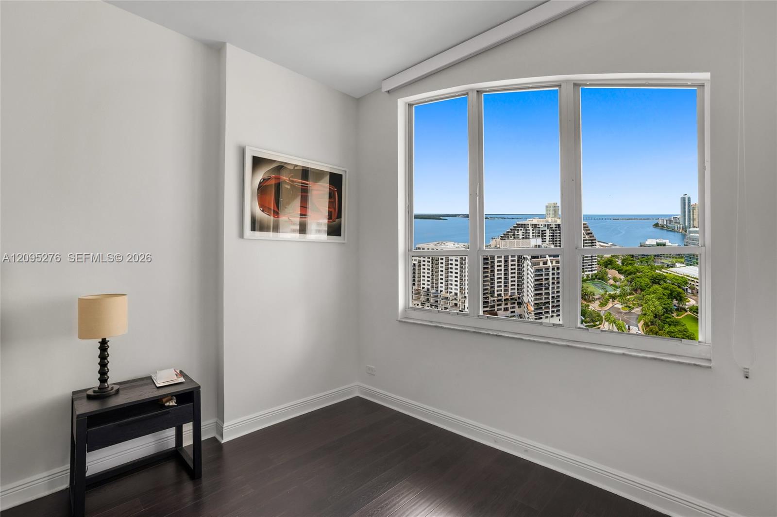 Bright high-rise room with dark wood flooring and a large window overlooking water, buildings, and greenery.