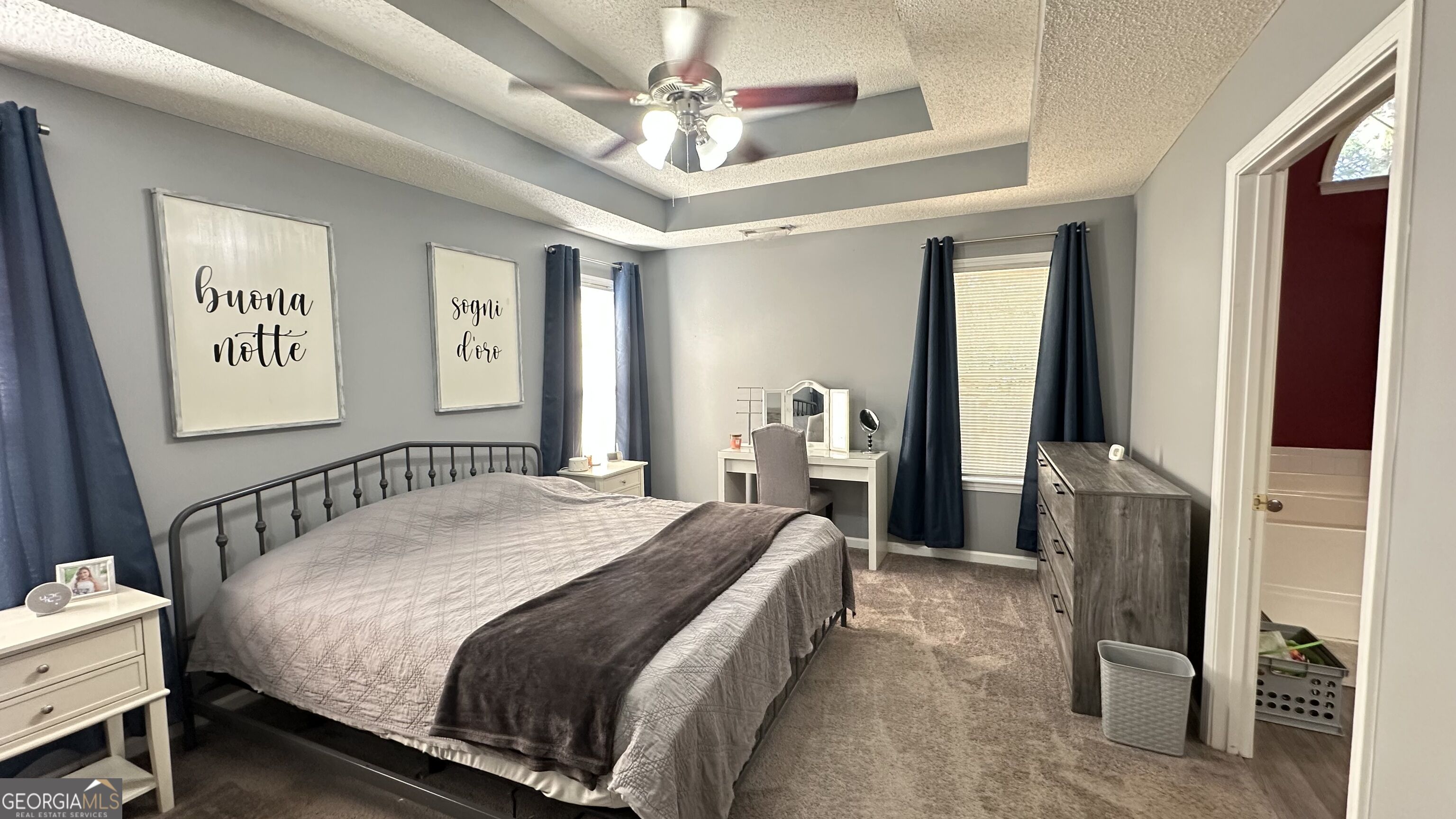 Furnished bedroom with a metal-frame bed, navy curtains, tray ceiling, vanity, and dresser.