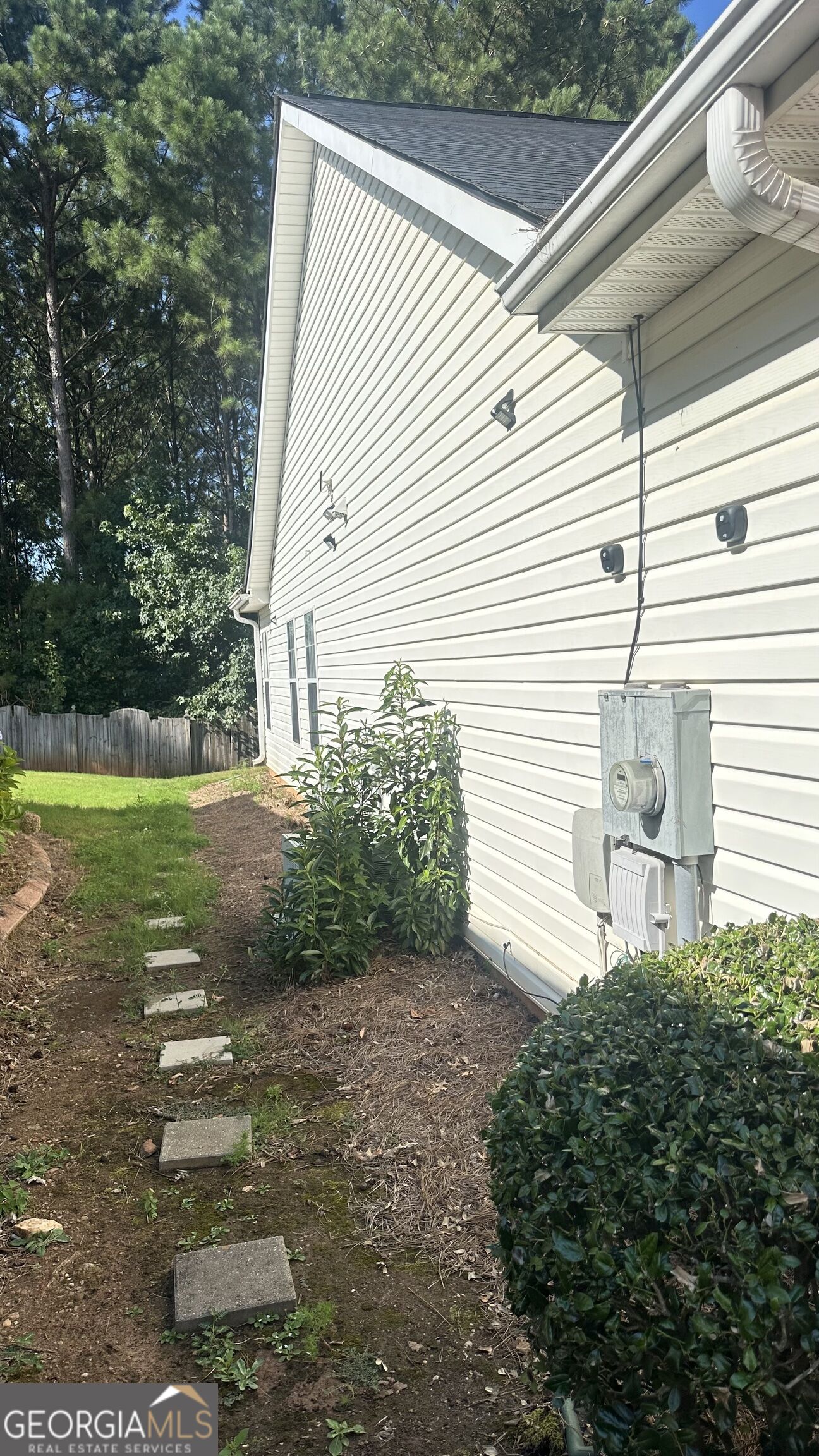 House side with vinyl siding, utility meter, stepping stones, and shrubs along a narrow yard.
