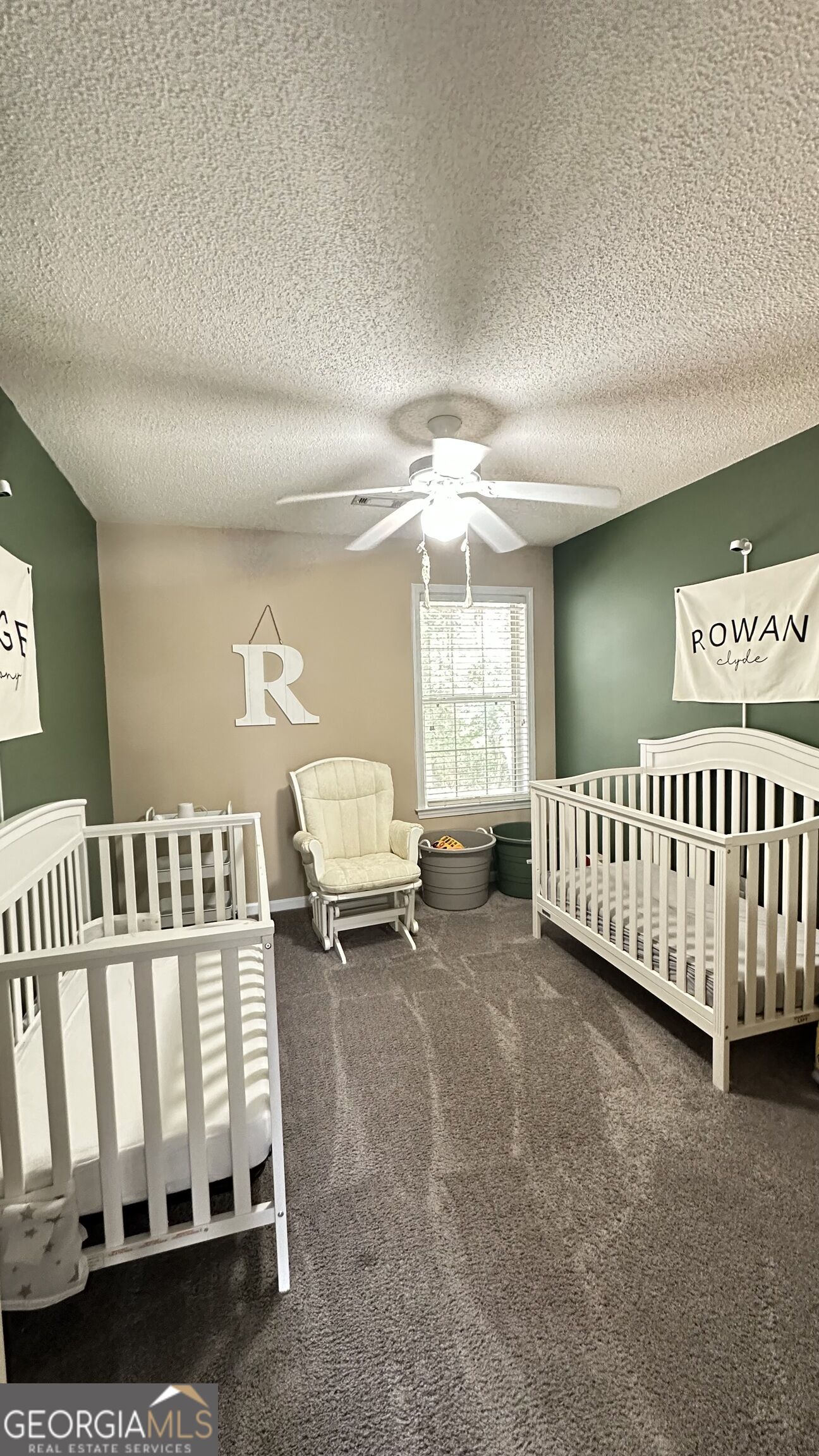 Furnished nursery with two white cribs, a cream rocking chair, green accent walls, and a ceiling fan.