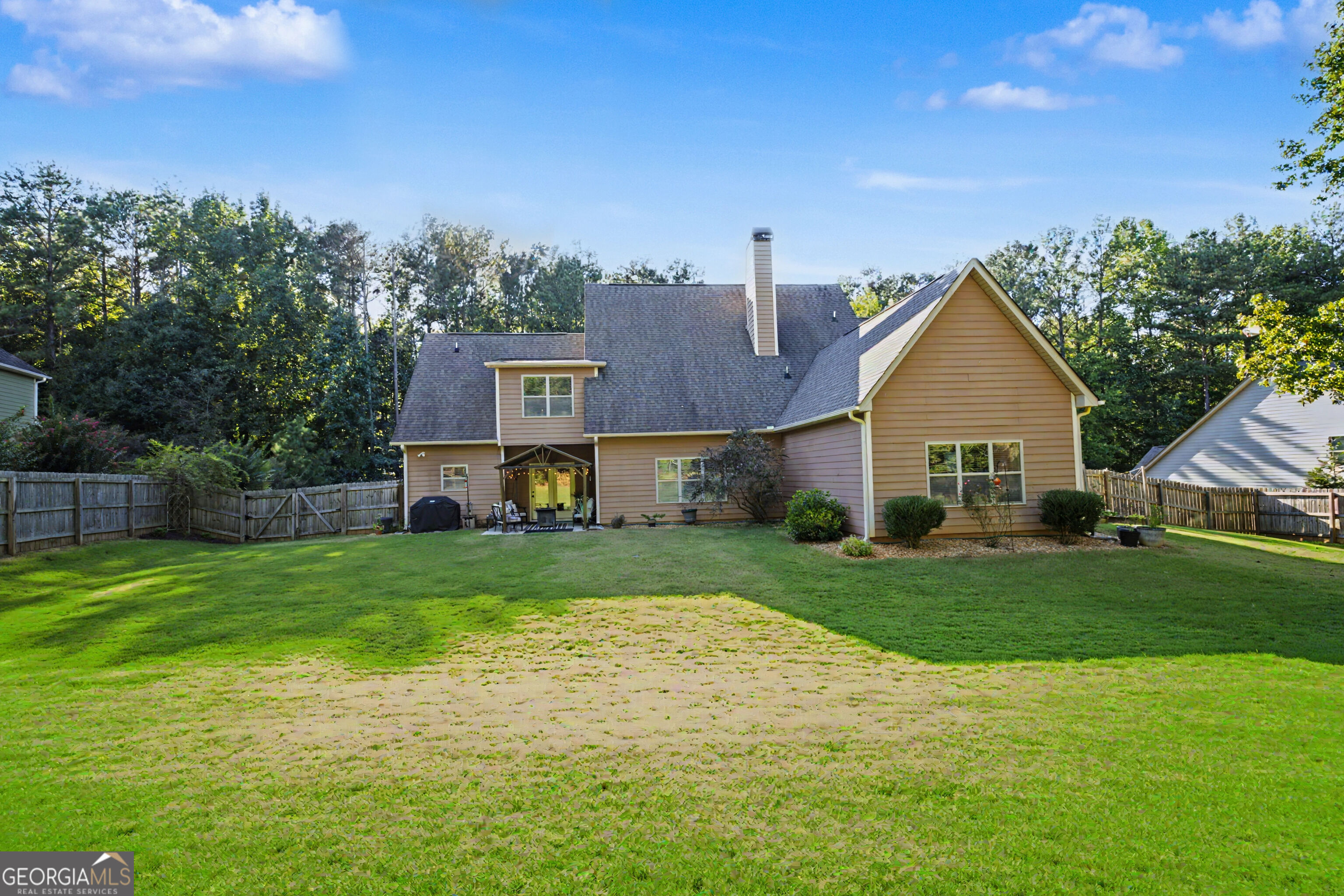 Rear of a tan two-story house with a fenced lawn, patio seating, and mature trees.