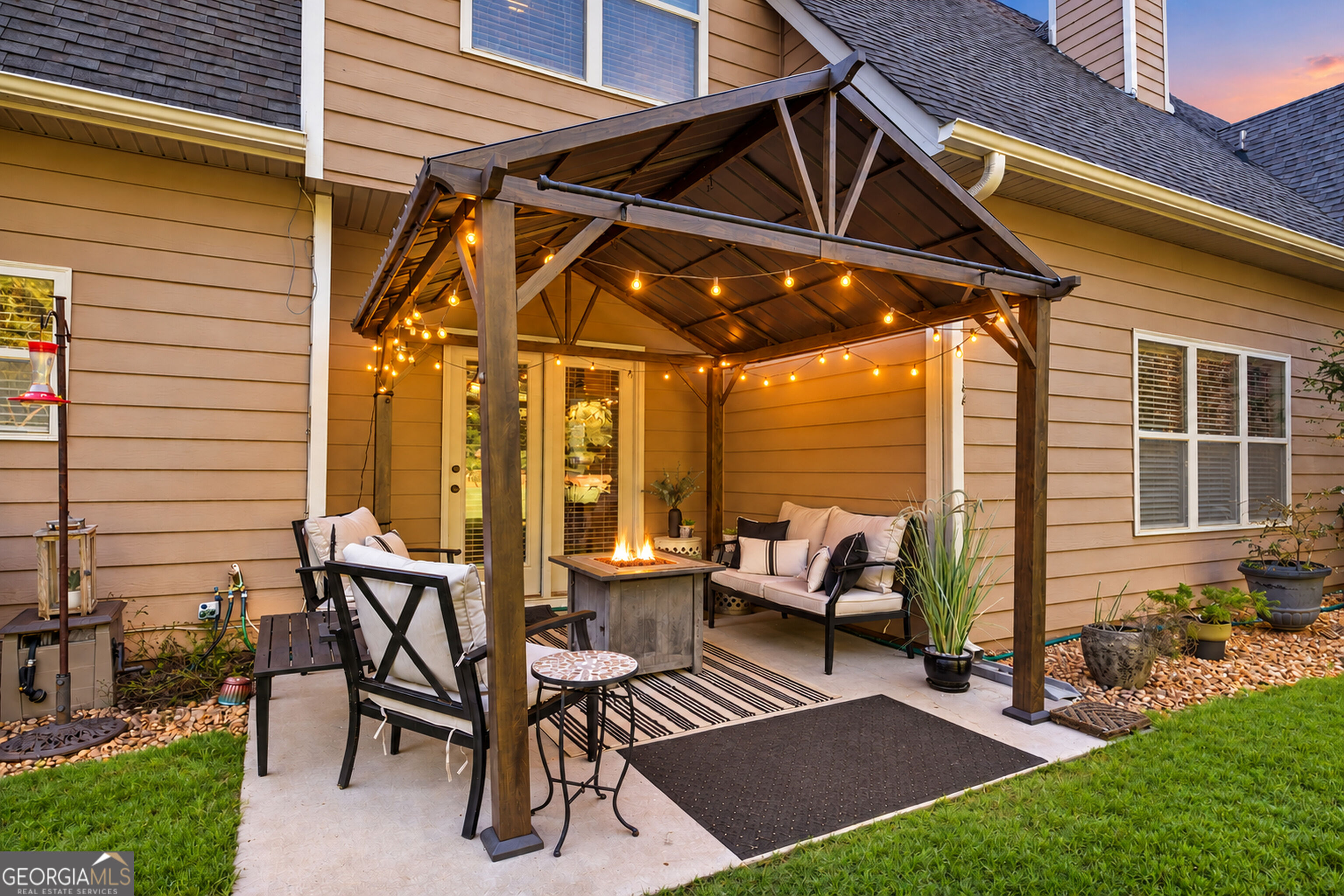 Backyard patio with a light-strung wooden pergola, cushioned seating, and a fire table.