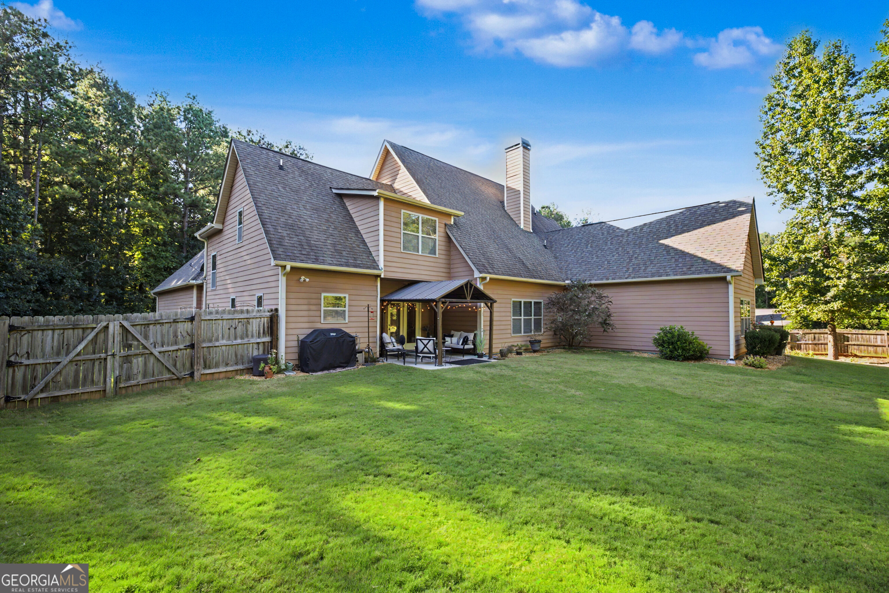 Rear of a tan two-story home with a covered, furnished patio, broad lawn, and wooden privacy fence.