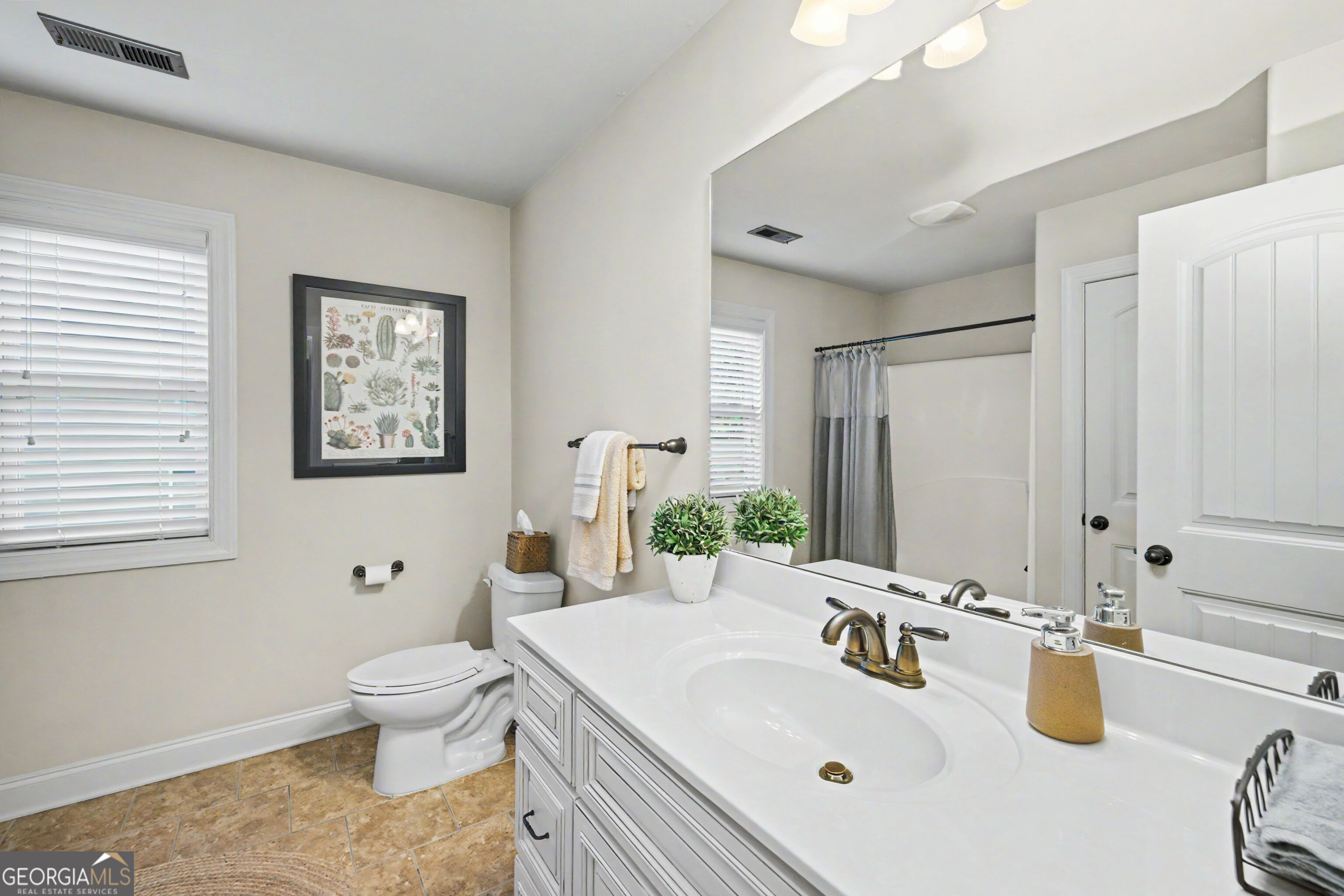 Bathroom with a white vanity, tan tile floor, botanical wall art, and gray shower curtain.