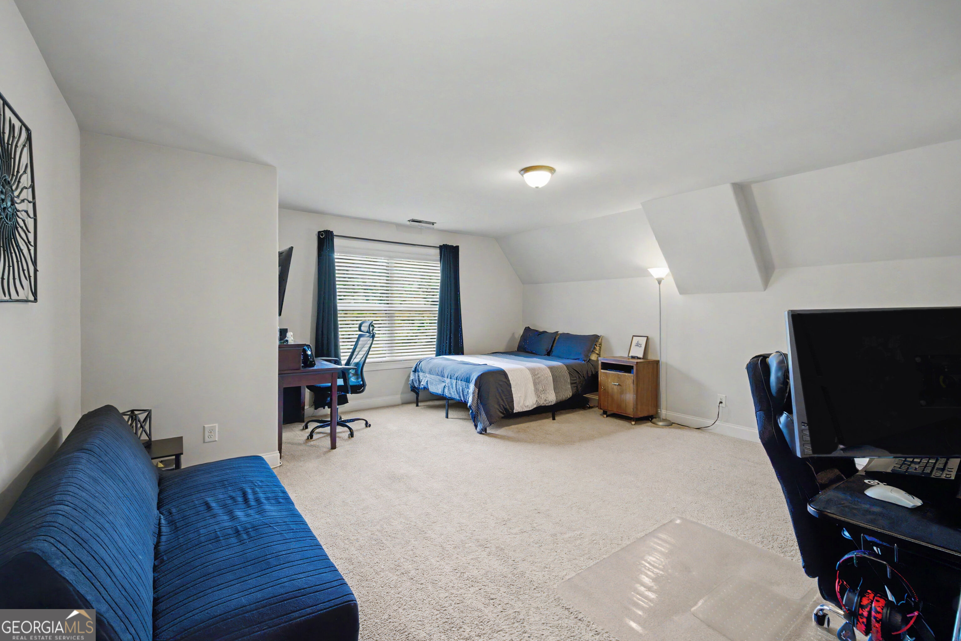 Furnished bedroom with a bed, blue sofa, desk, and computer setup beneath a sloped ceiling.