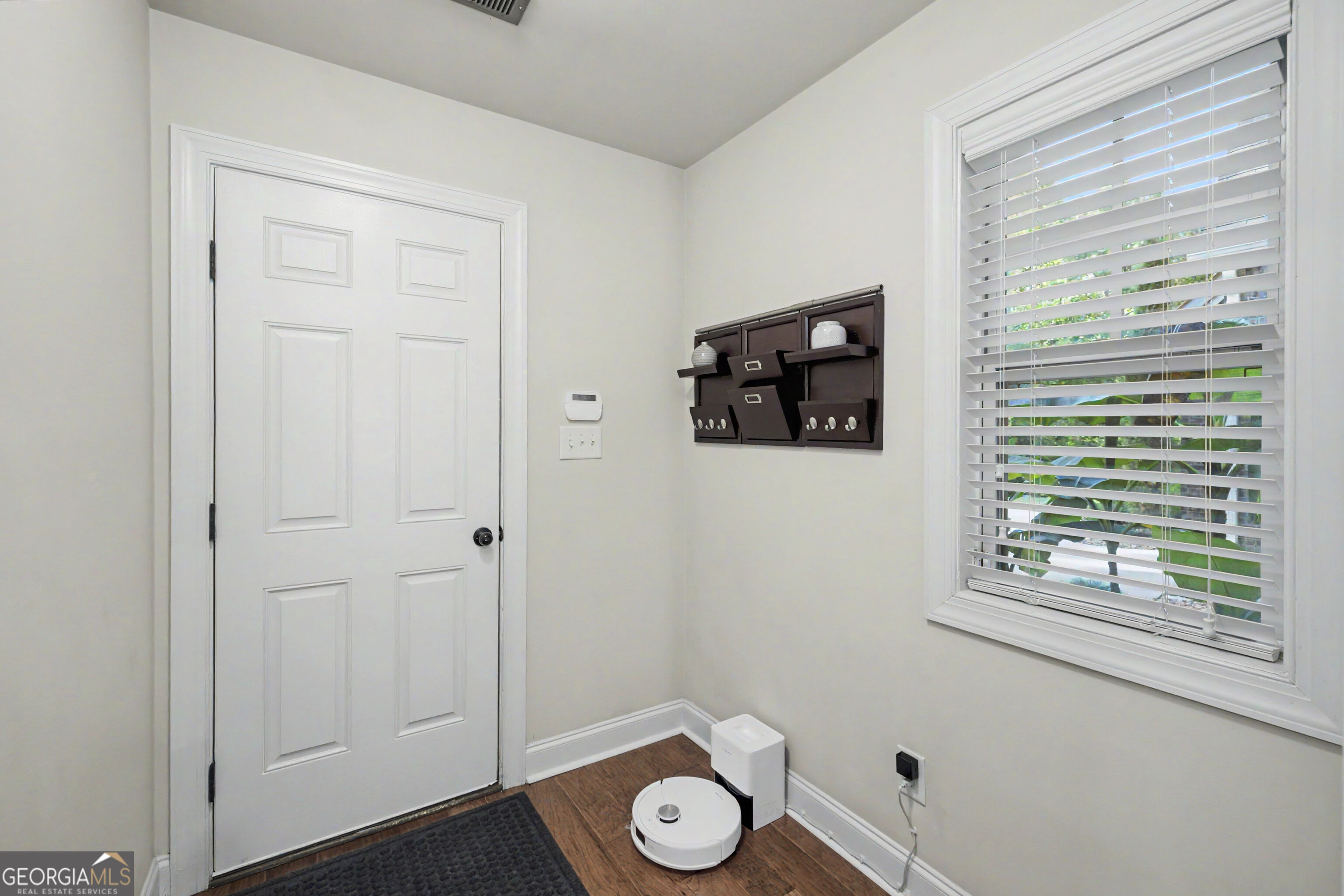 Entry foyer with a white paneled front door, window blinds, wall organizer, and robot vacuum.