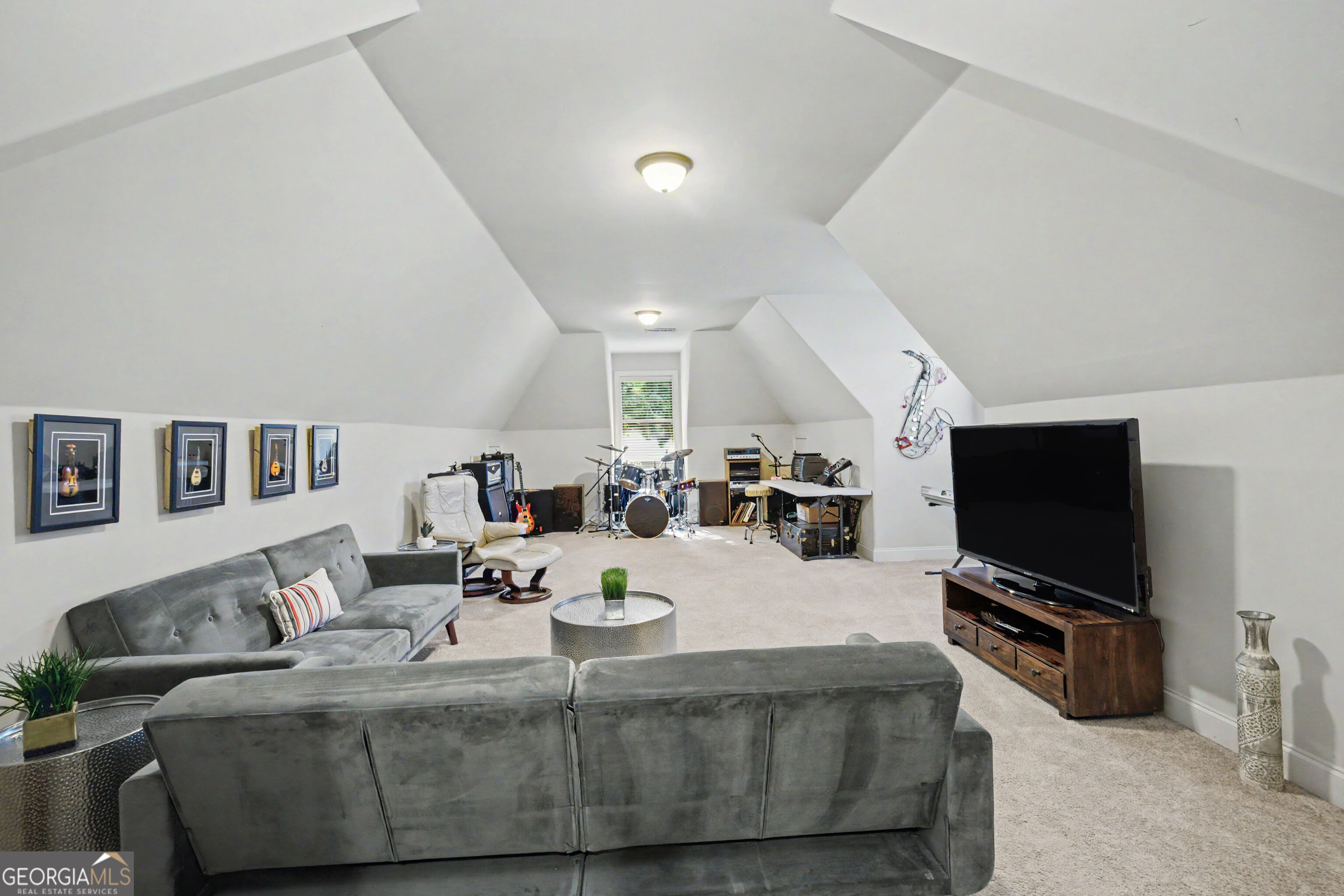Furnished vaulted bonus room with gray sofas, a television, and a drum kit and guitars.