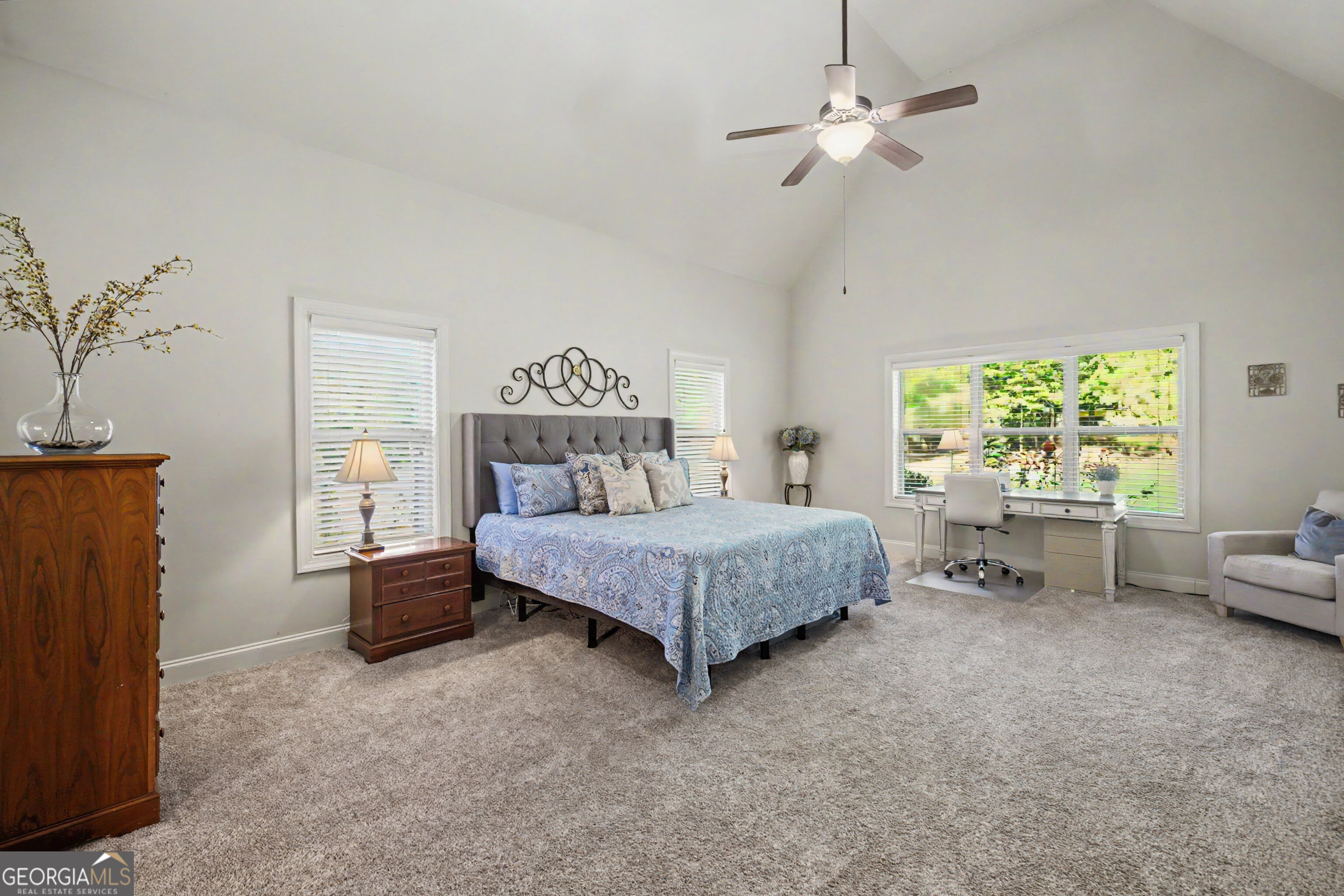 Bedroom with a vaulted ceiling, blue patterned bedding, and a desk beside a wide window.