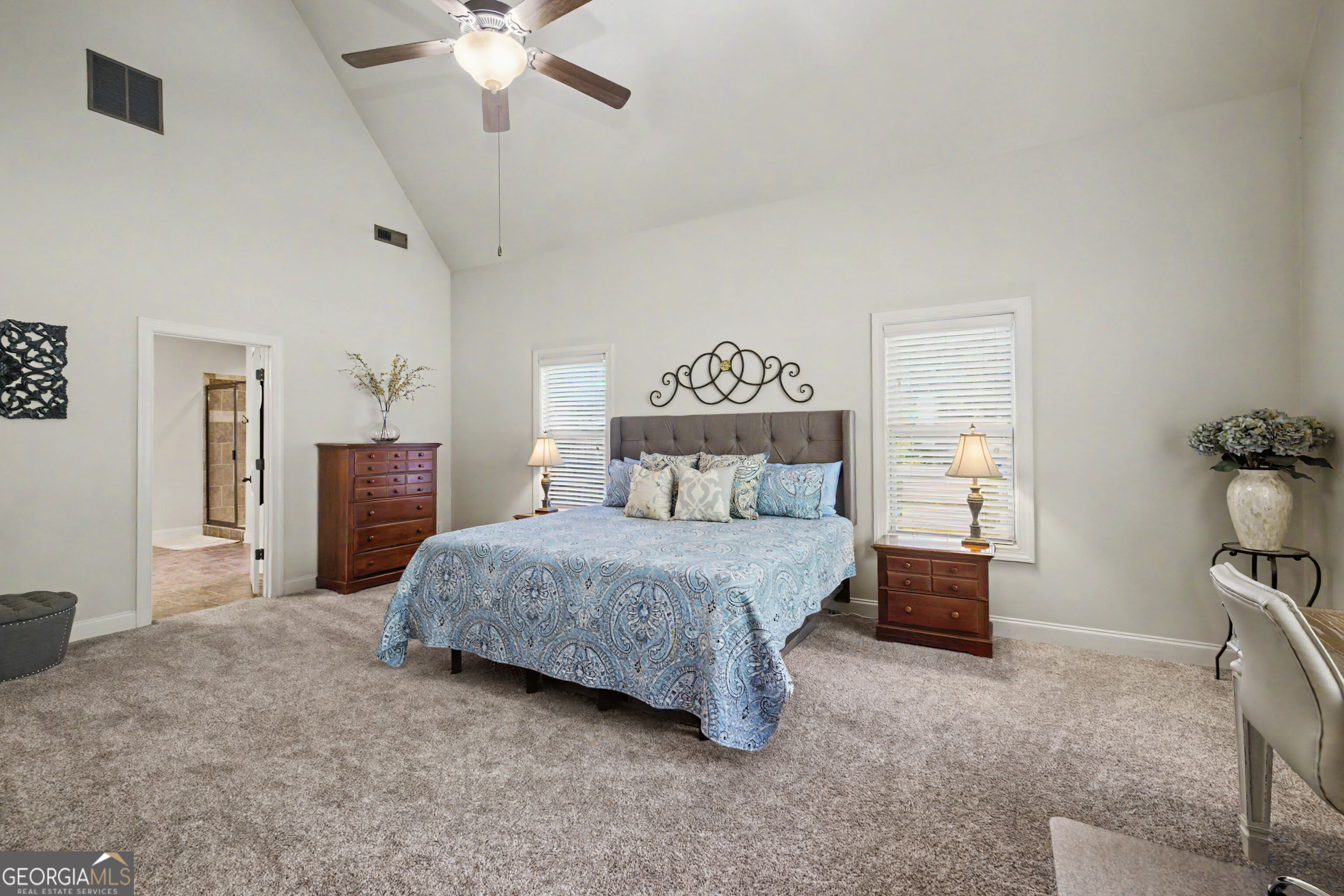 Staged bedroom with a vaulted ceiling, blue patterned bedding, and an open doorway to a tiled bathroom.