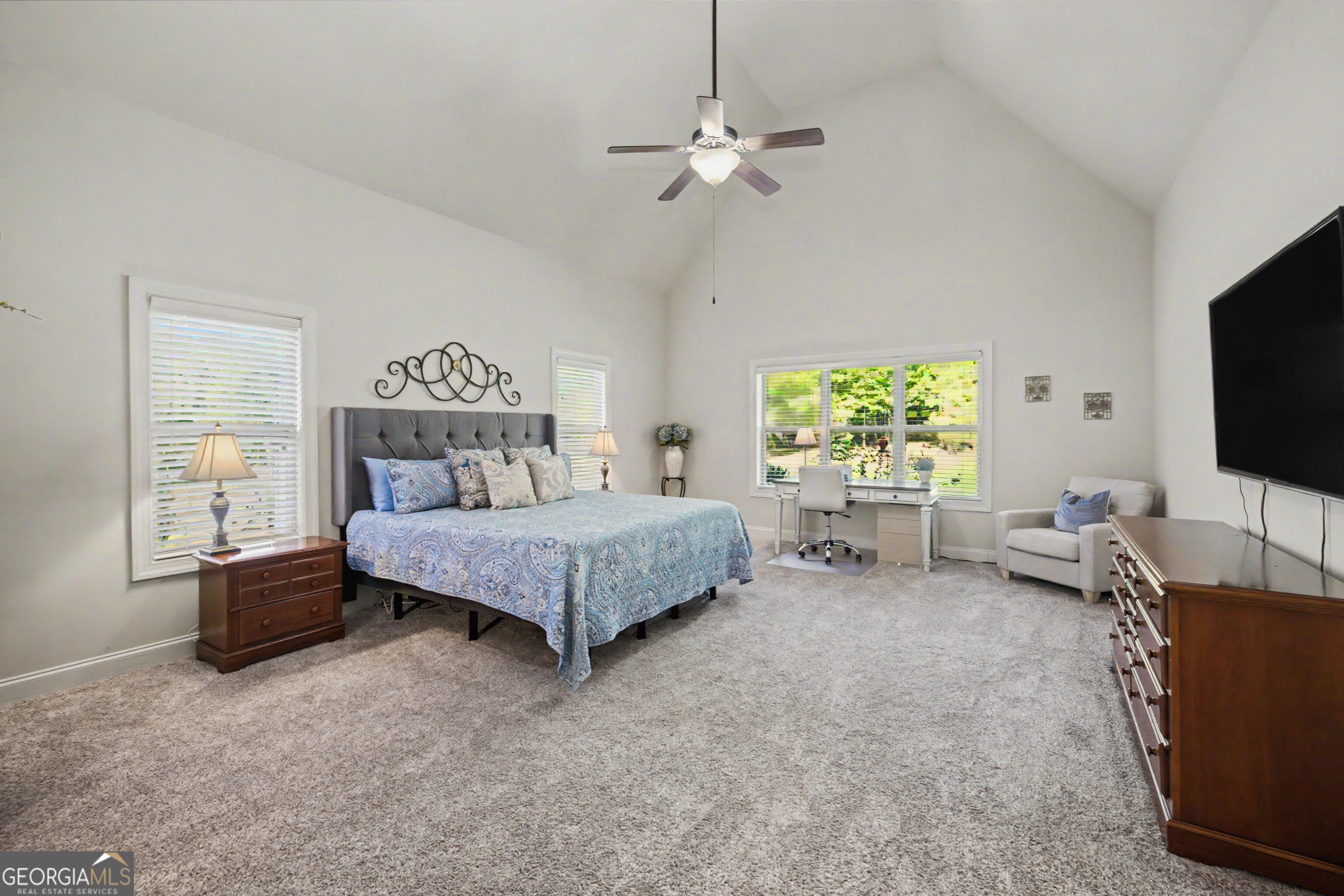 Furnished bedroom with a vaulted ceiling, blue patterned bedspread, desk by the window, and carpeted floor.