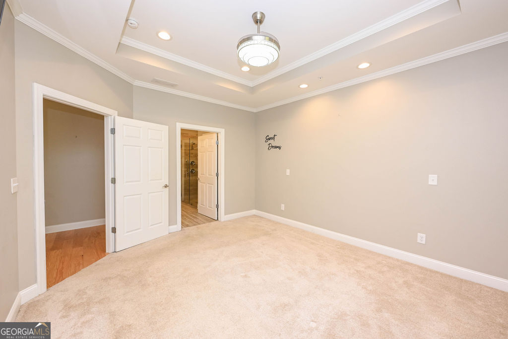Empty bedroom with beige carpet, tray ceiling, and open doors to an adjoining bathroom.