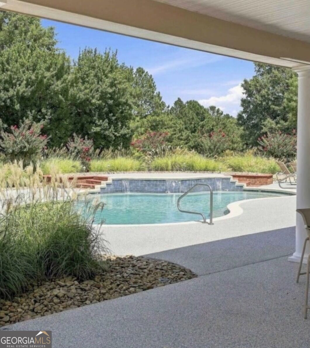 Outdoor pool with a curved deck, raised tiled wall, and landscaped greenery, viewed from a covered patio.