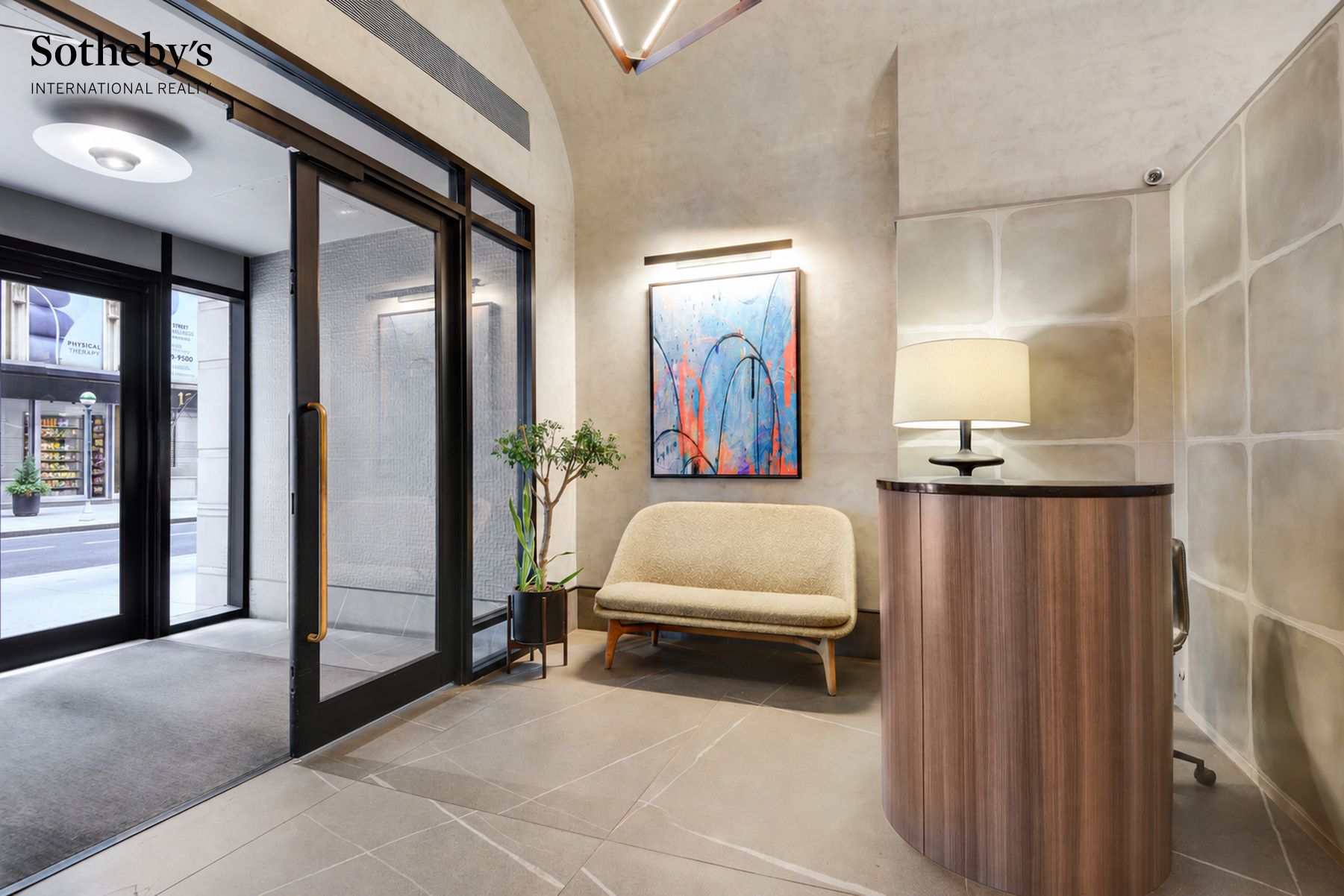 Building lobby with a curved wood reception desk, cream settee, abstract art, and glass entrance doors.
