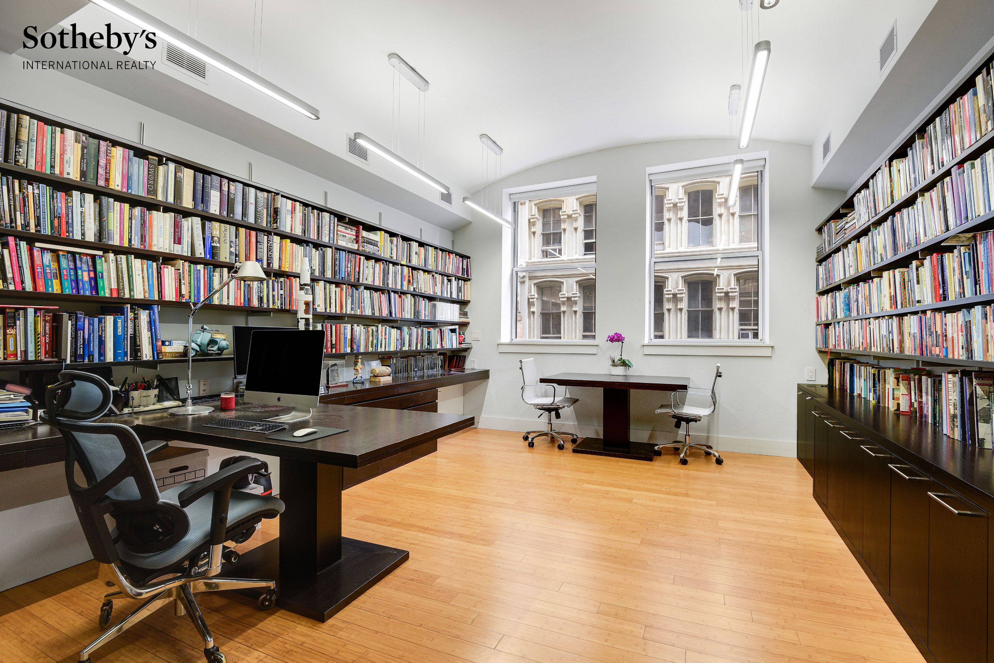 Home office with floor-to-ceiling bookshelves, dark wood desks, and two tall windows.
