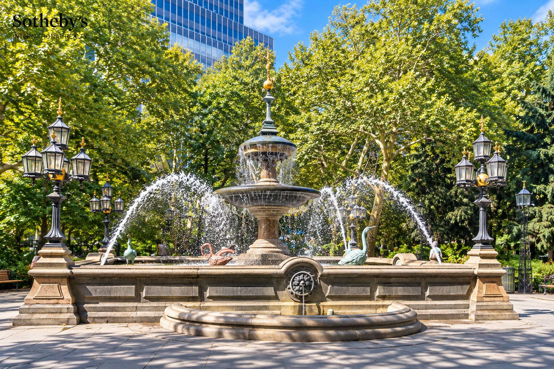 Stone park fountain spraying water beneath leafy trees and ornate street lamps.