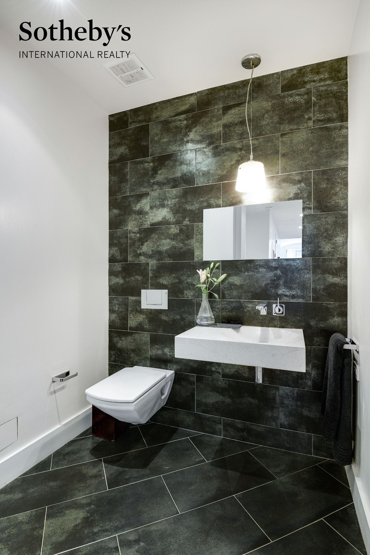 Bathroom with dark stone tile, a floating white sink, wall-mounted toilet, and pendant light.