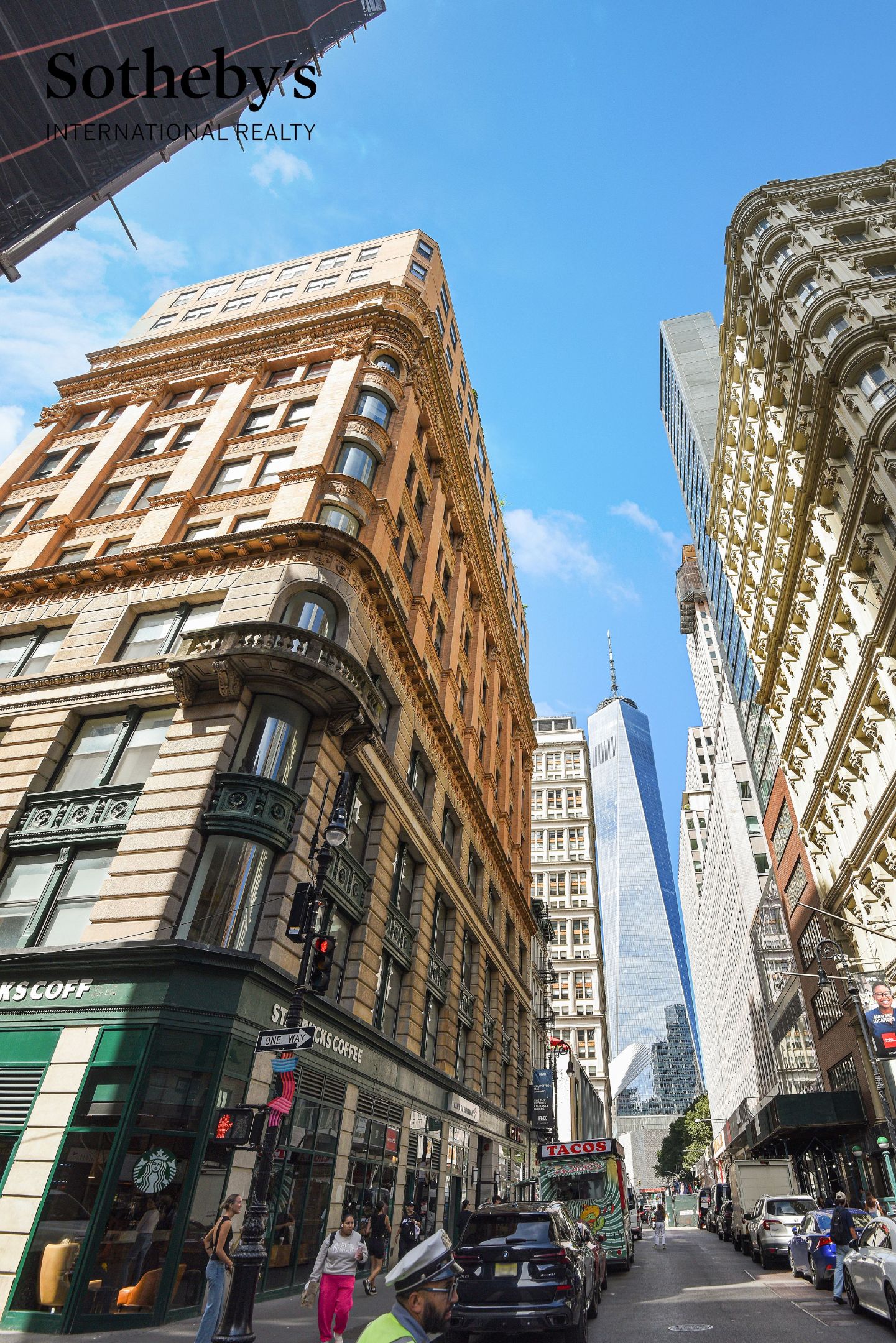 Lower Manhattan street lined with ornate buildings, with One World Trade Center in the distance.