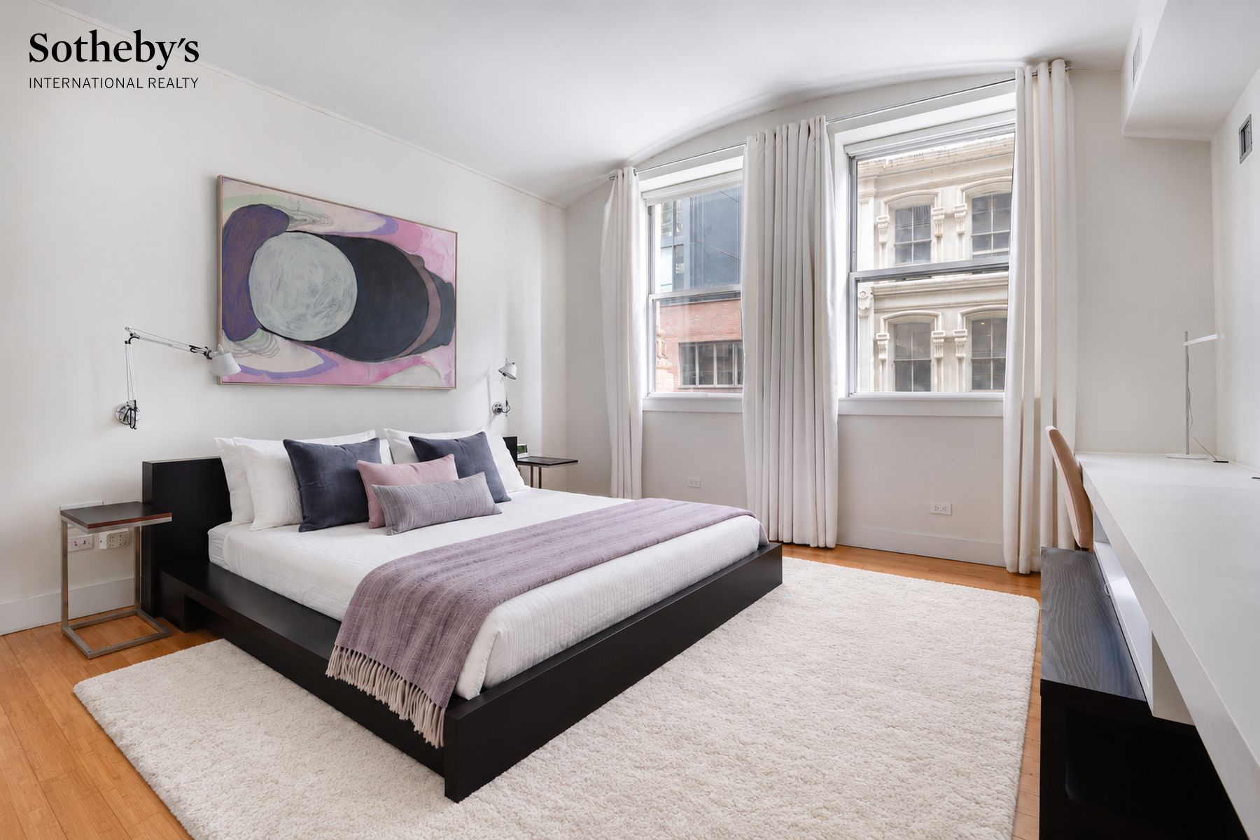 Staged bedroom with a black platform bed, abstract artwork, and tall city-facing windows.