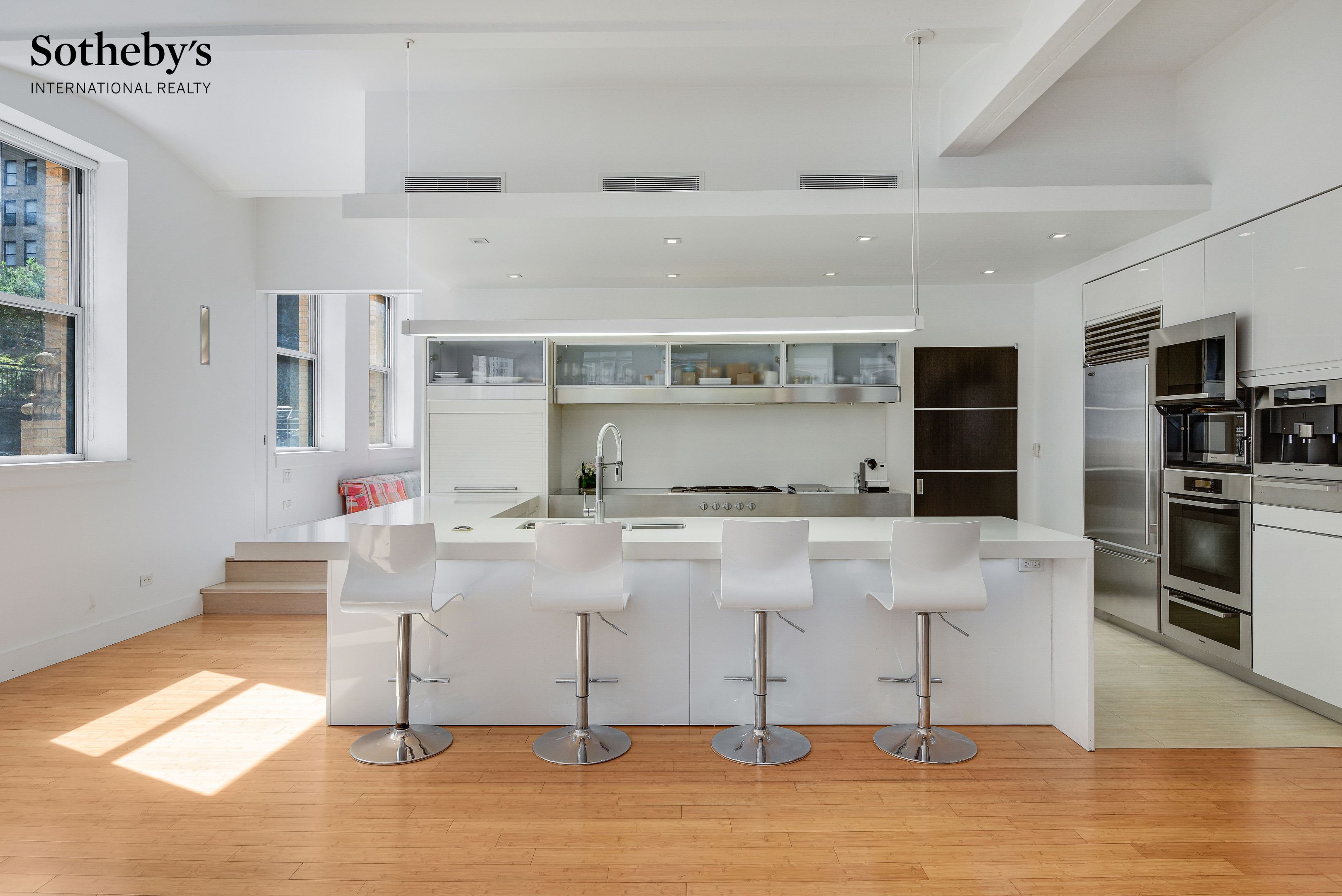 Modern white kitchen with a large island, four bar stools, and stainless steel appliances.