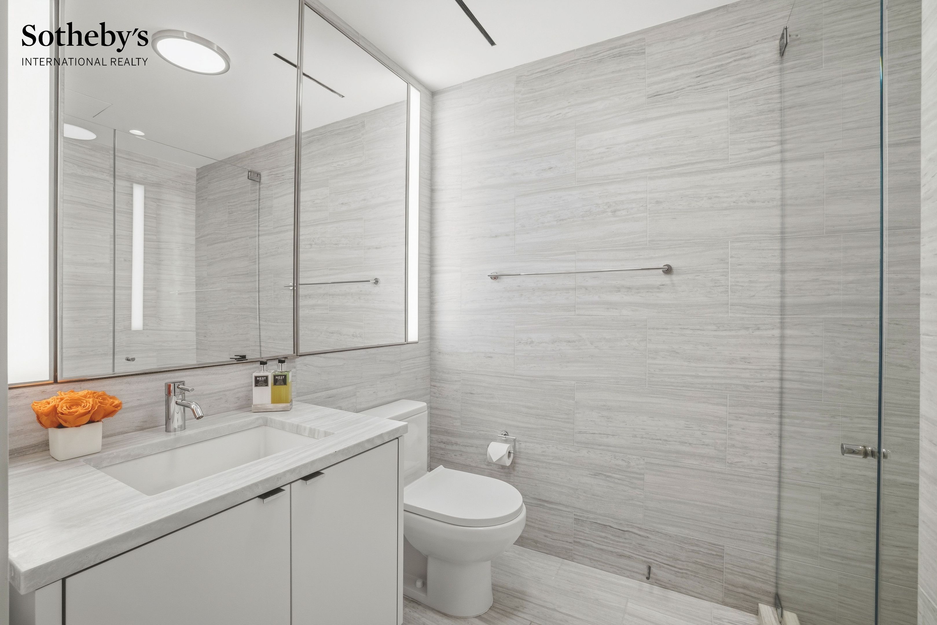 Modern bathroom with pale gray tile, a glass shower, and a white vanity topped with orange roses.