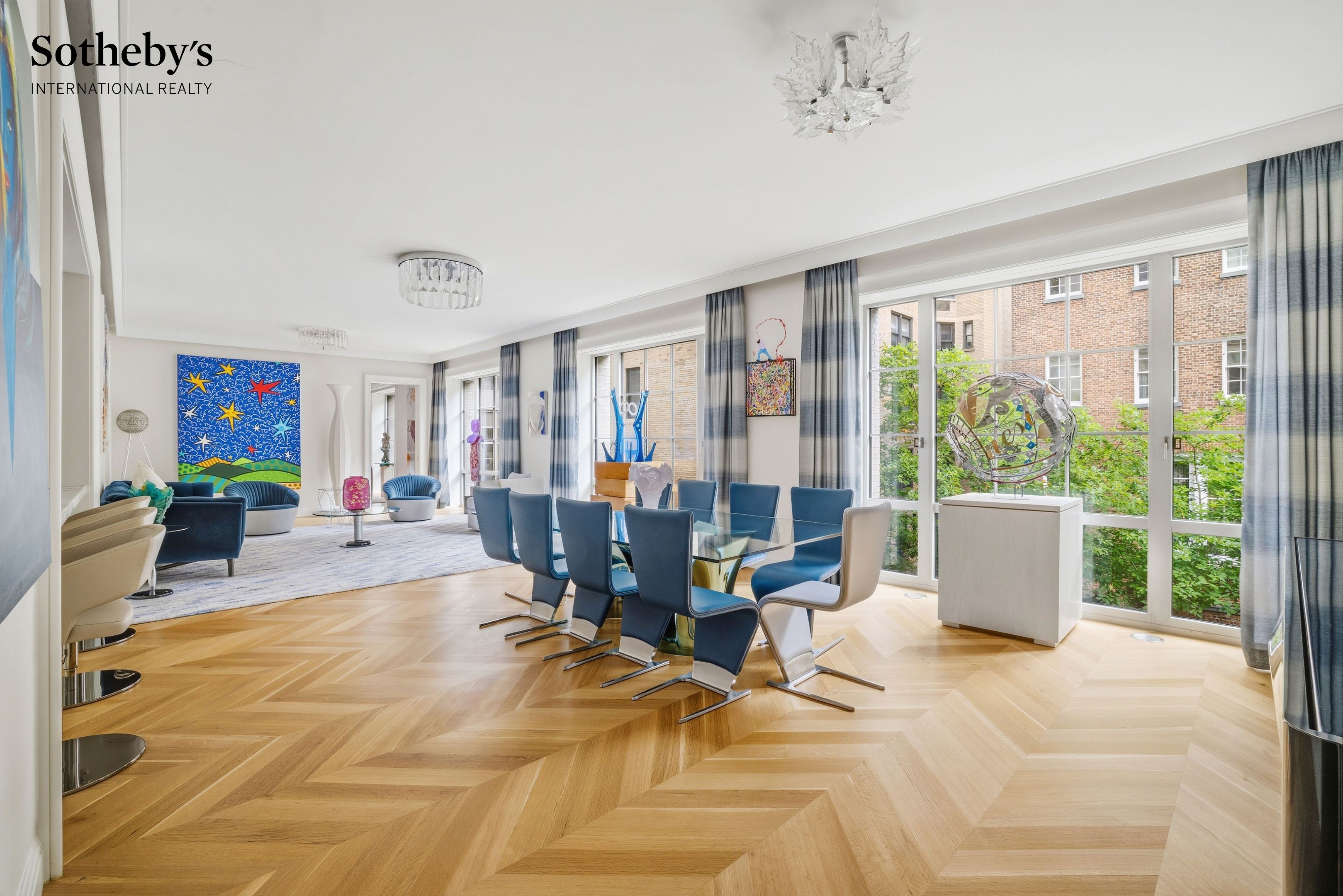 Living and dining room with blue chairs, colorful artwork, herringbone floors, and large windows.