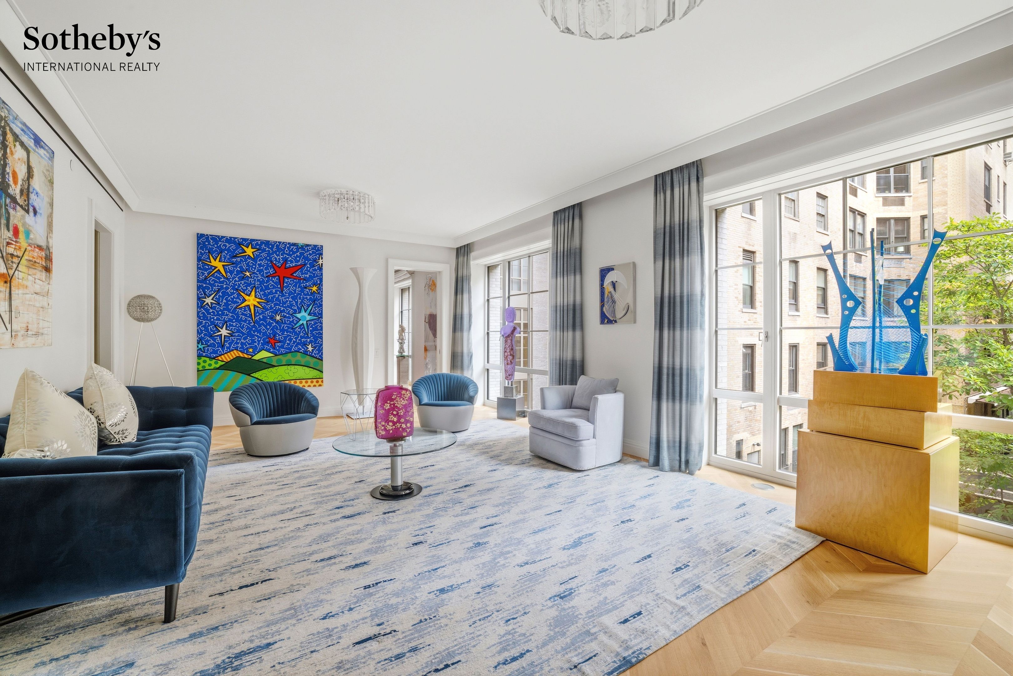 Living room with blue velvet seating, colorful artwork, and large windows overlooking a leafy courtyard.