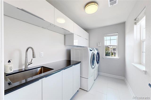 Laundry room with white cabinets, black countertop sink, front-loading washer and dryer, and large windows.