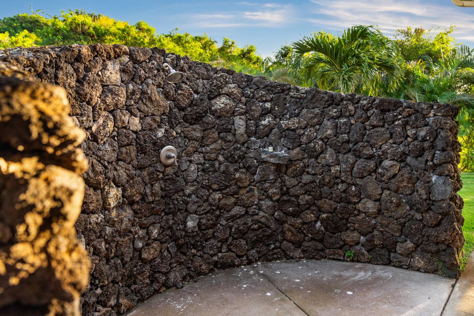 Outdoor shower enclosed by a rough lava-rock wall, with tropical palms visible above it.