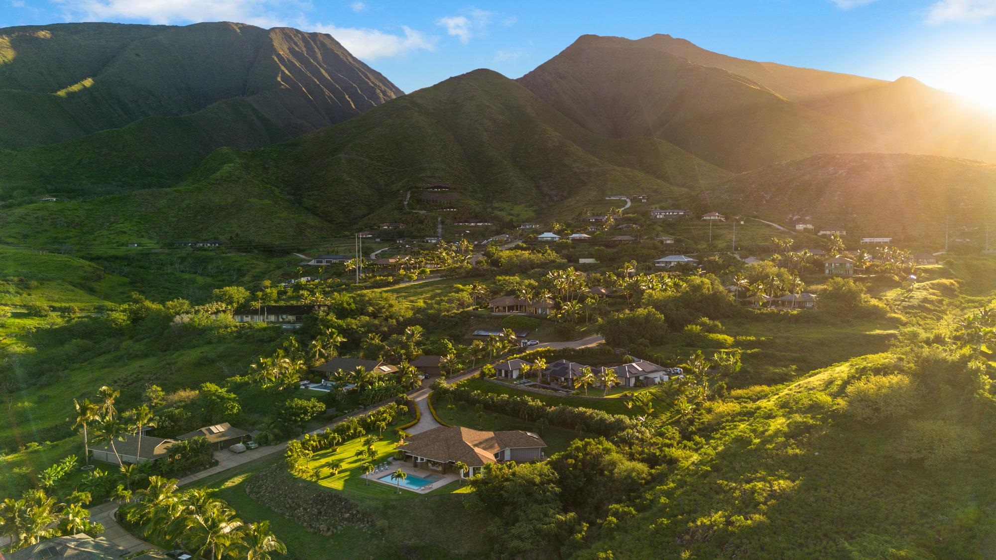 Aerial view of homes and palm trees beneath steep green mountains in golden-hour light.
