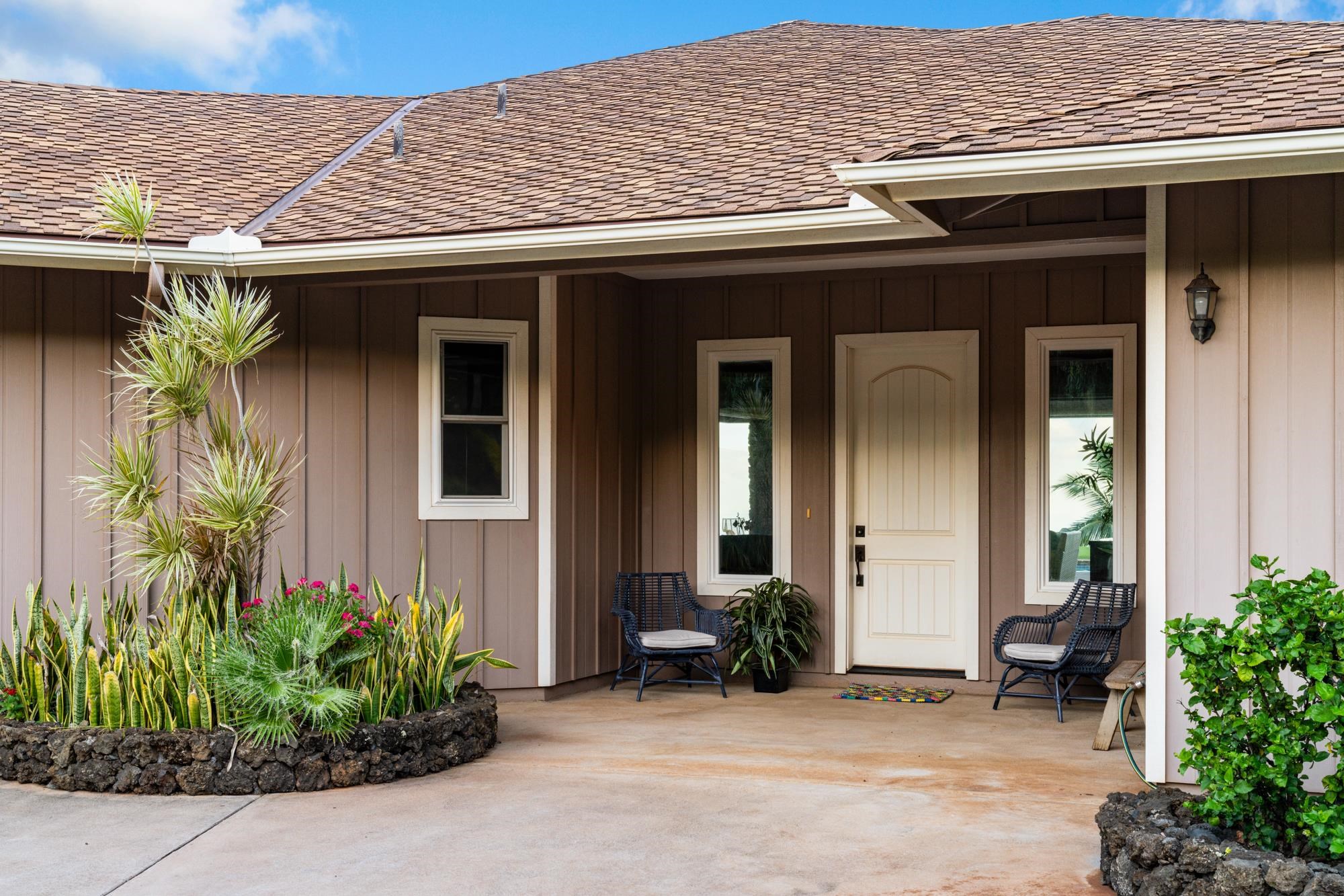 Home’s covered front porch with a light entry door, two chairs, and tropical landscaping.