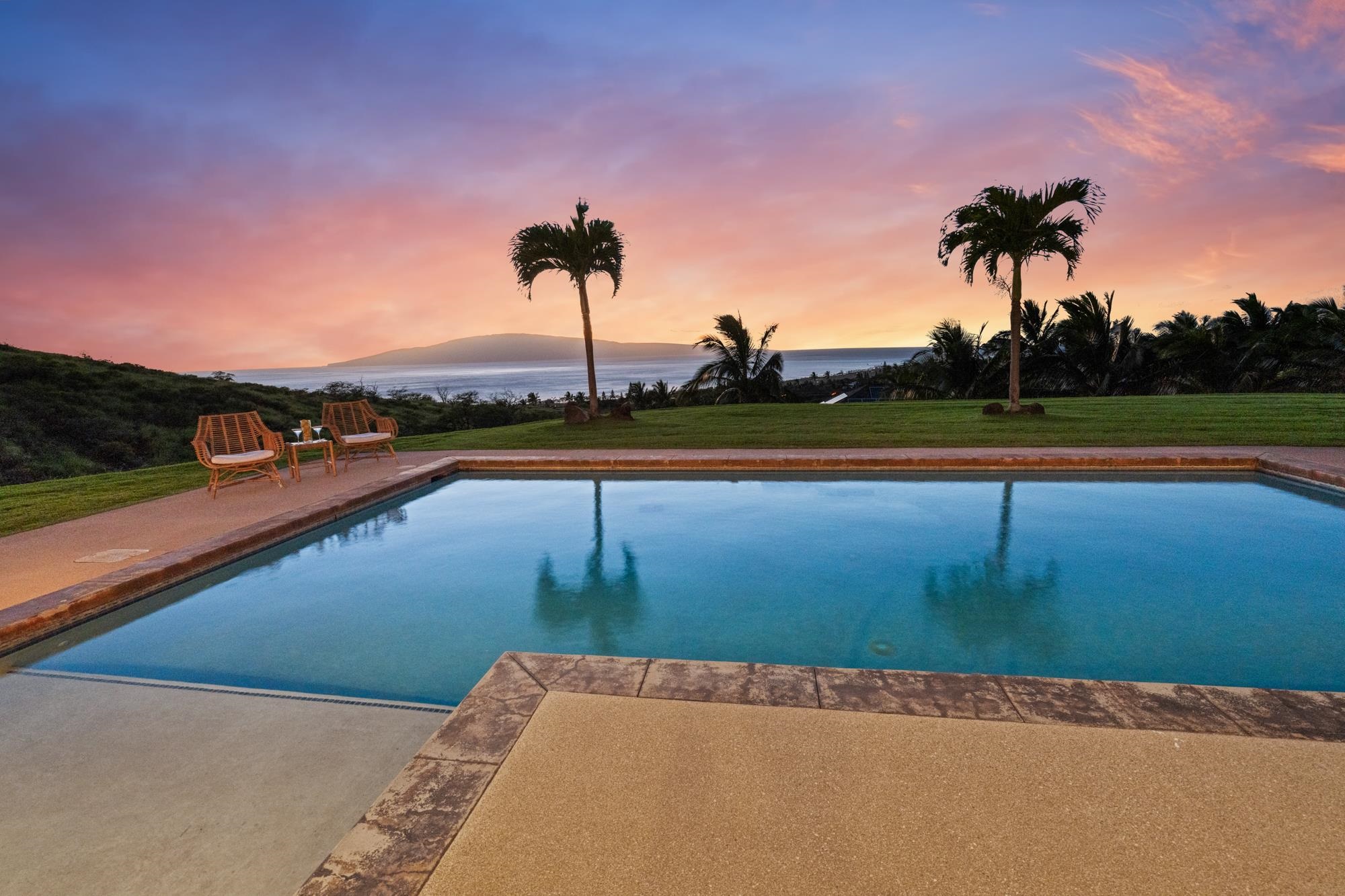 Swimming pool overlooking the ocean at sunset, with palm trees and lounge chairs.
