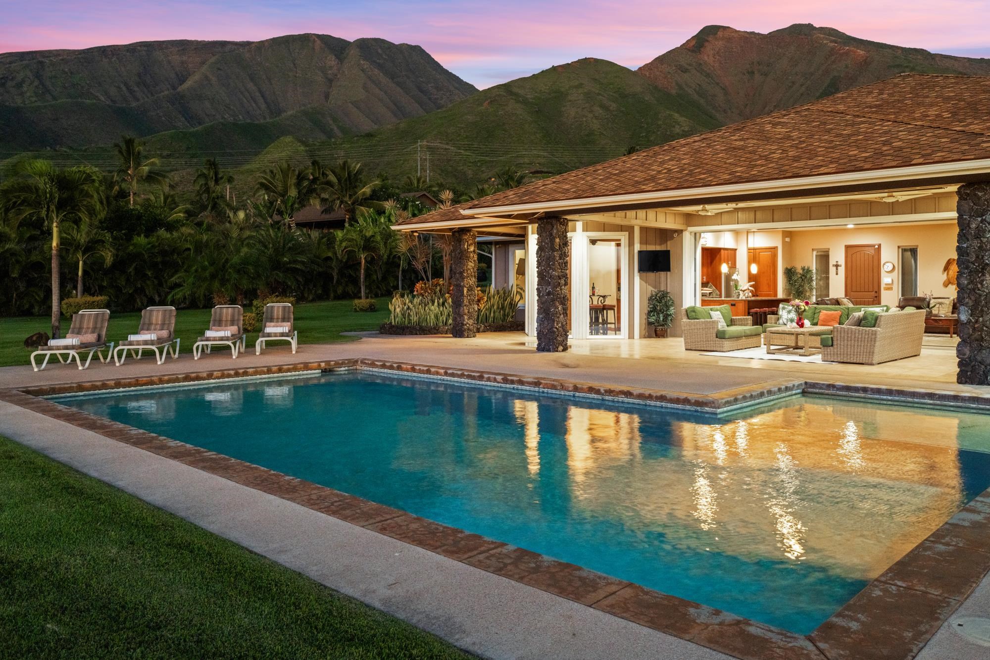 Mountain-view home with a lit lanai, tropical landscaping, and a swimming pool at dusk.