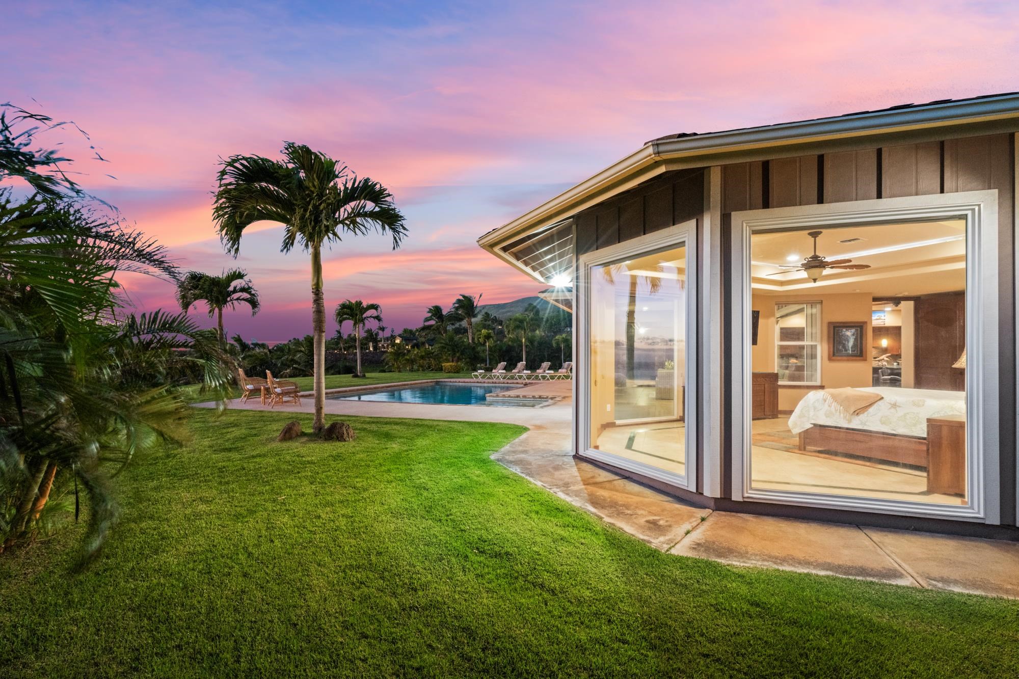 Home with a pool and palm-lined lawn at dusk, viewed through large bedroom windows.