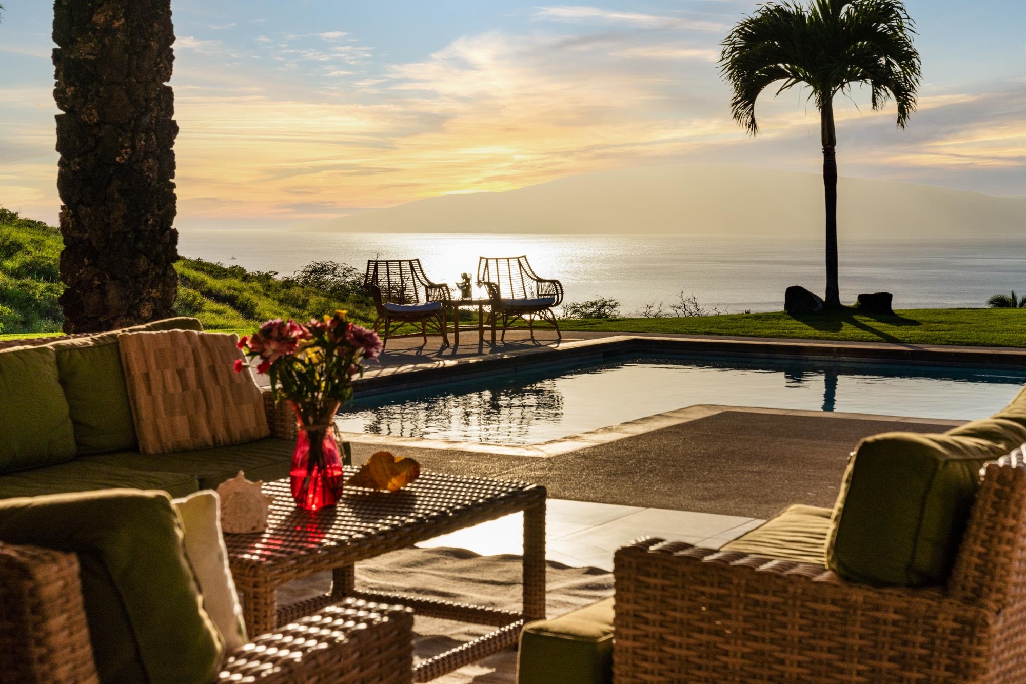 Oceanfront pool patio with wicker seating, palm trees, and a distant island at sunset.