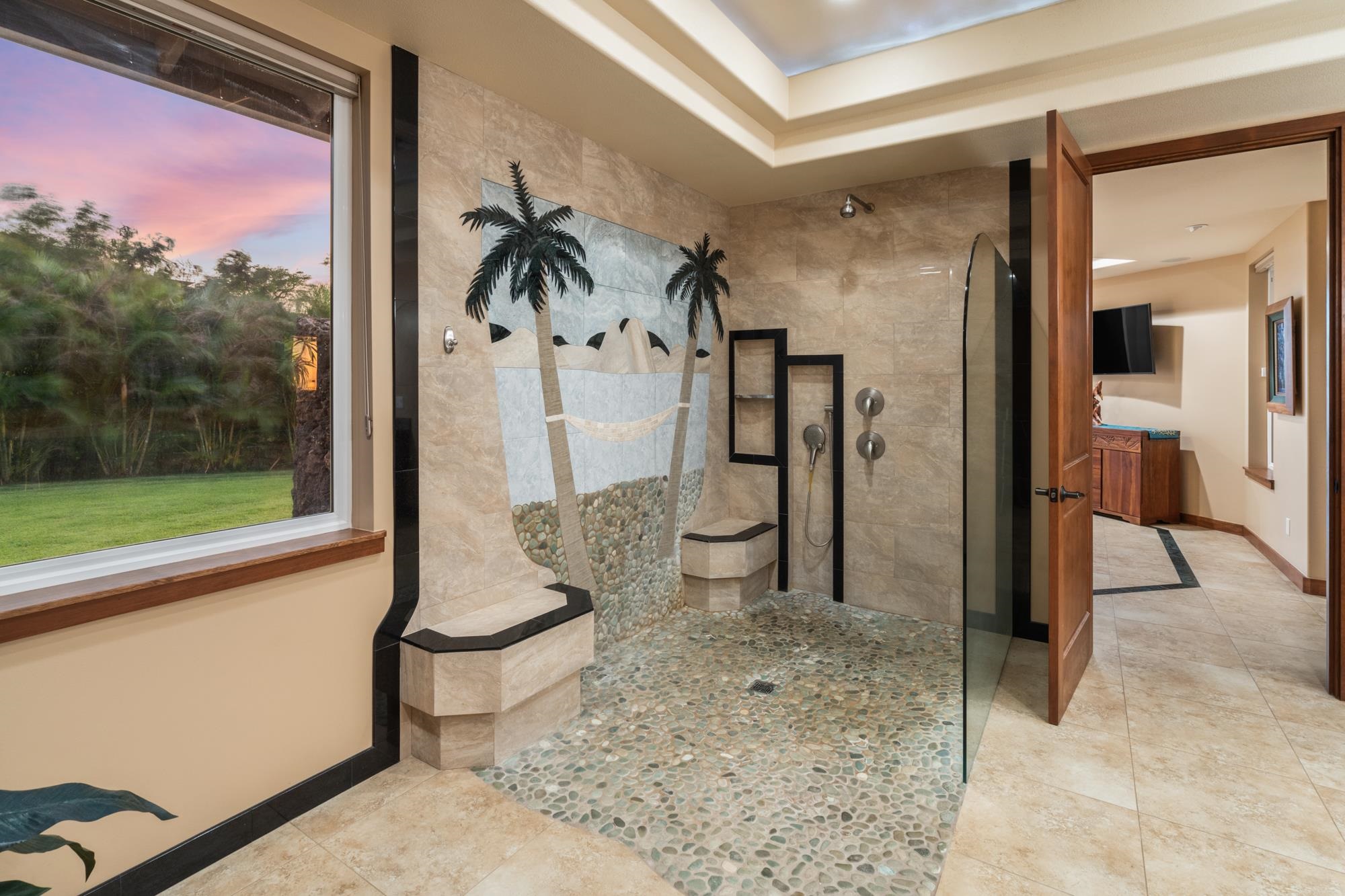 Bathroom with a pebble-floor walk-in shower, built-in bench, and palm-tree tile mosaic beside a large window.