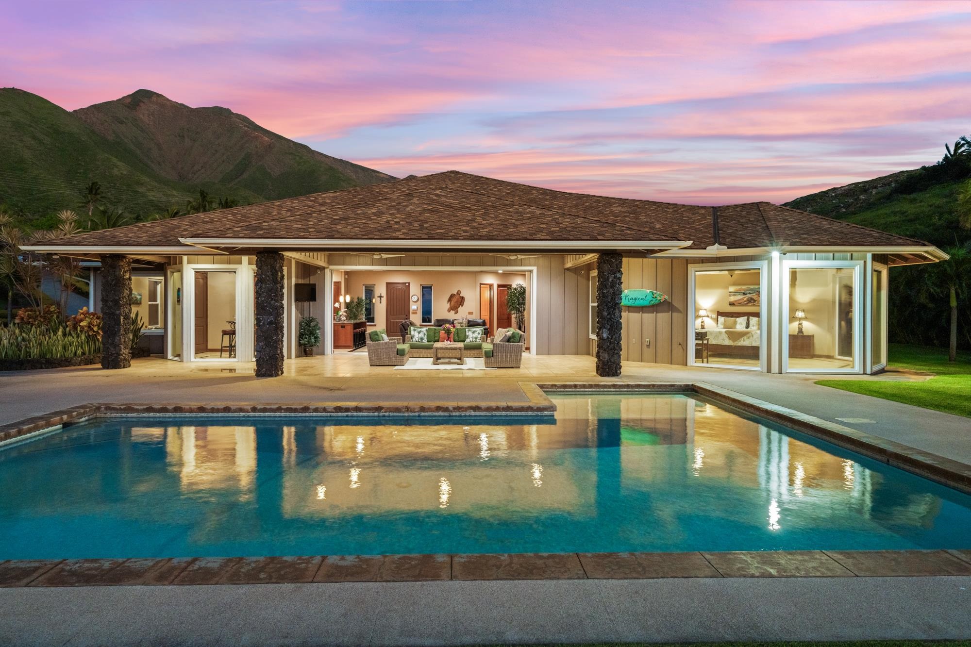 Single-story home and covered lanai reflected in a swimming pool, with mountains beneath a pink dusk sky.