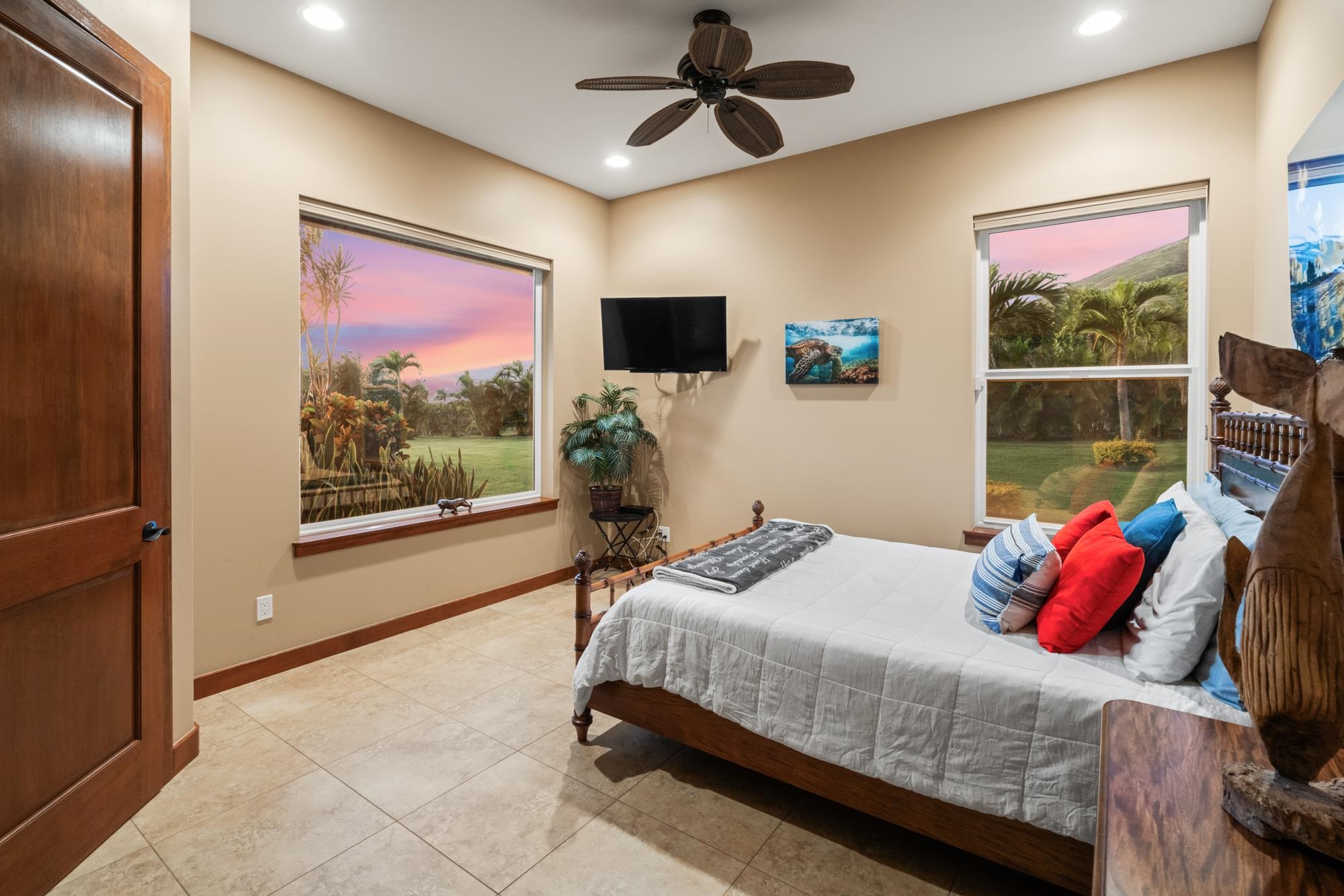 Bedroom with a wooden bed, colorful pillows, and windows overlooking tropical landscaping.