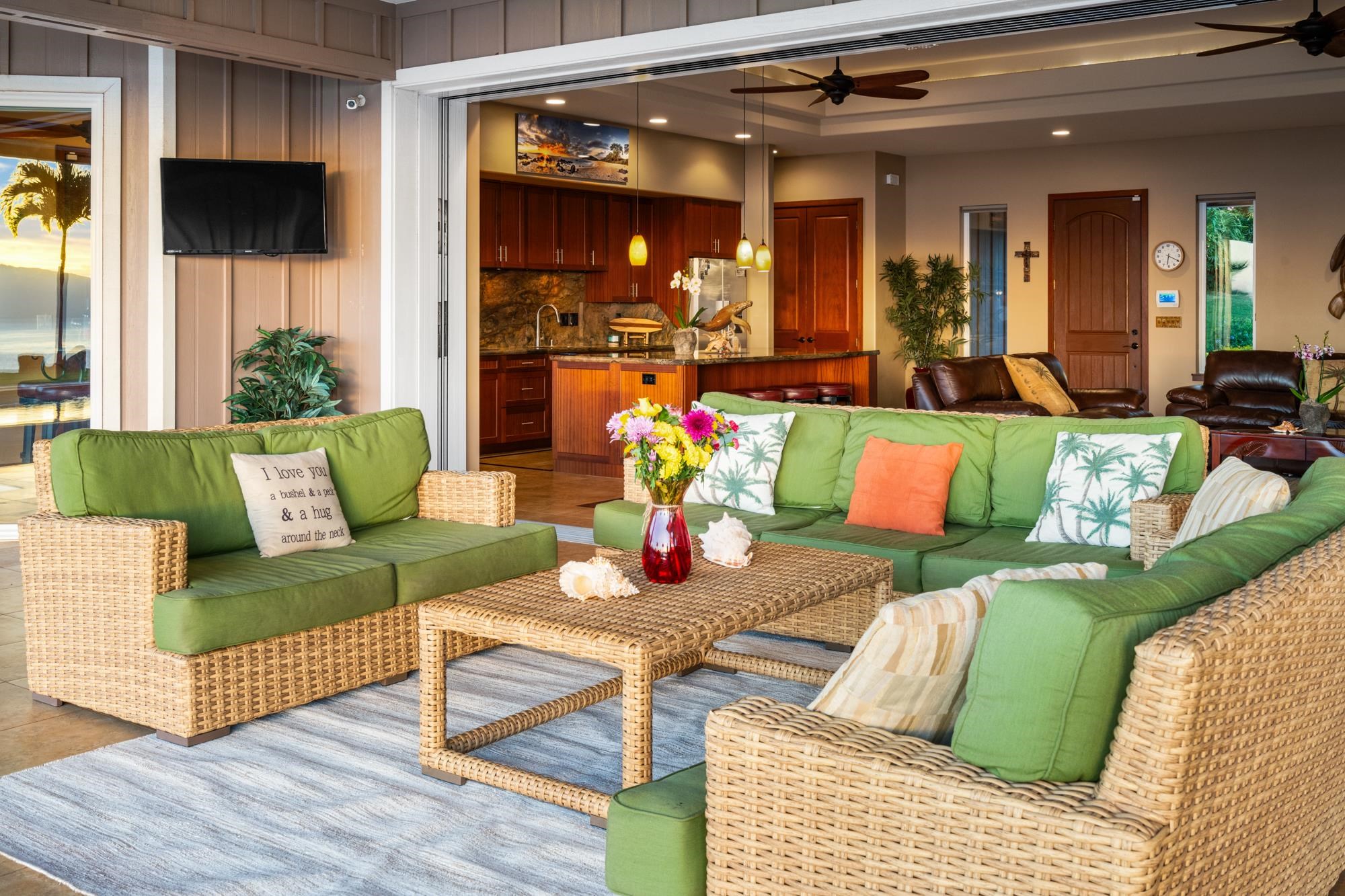 Open-plan living room with green wicker seating, woven coffee table, and wood-paneled kitchen beside ocean-view doors.