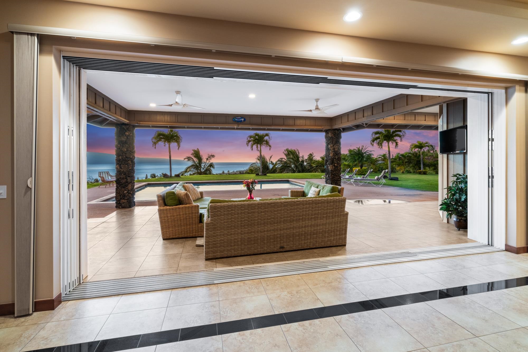 Furnished lanai opens to a pool and ocean view framed by palms at dusk.