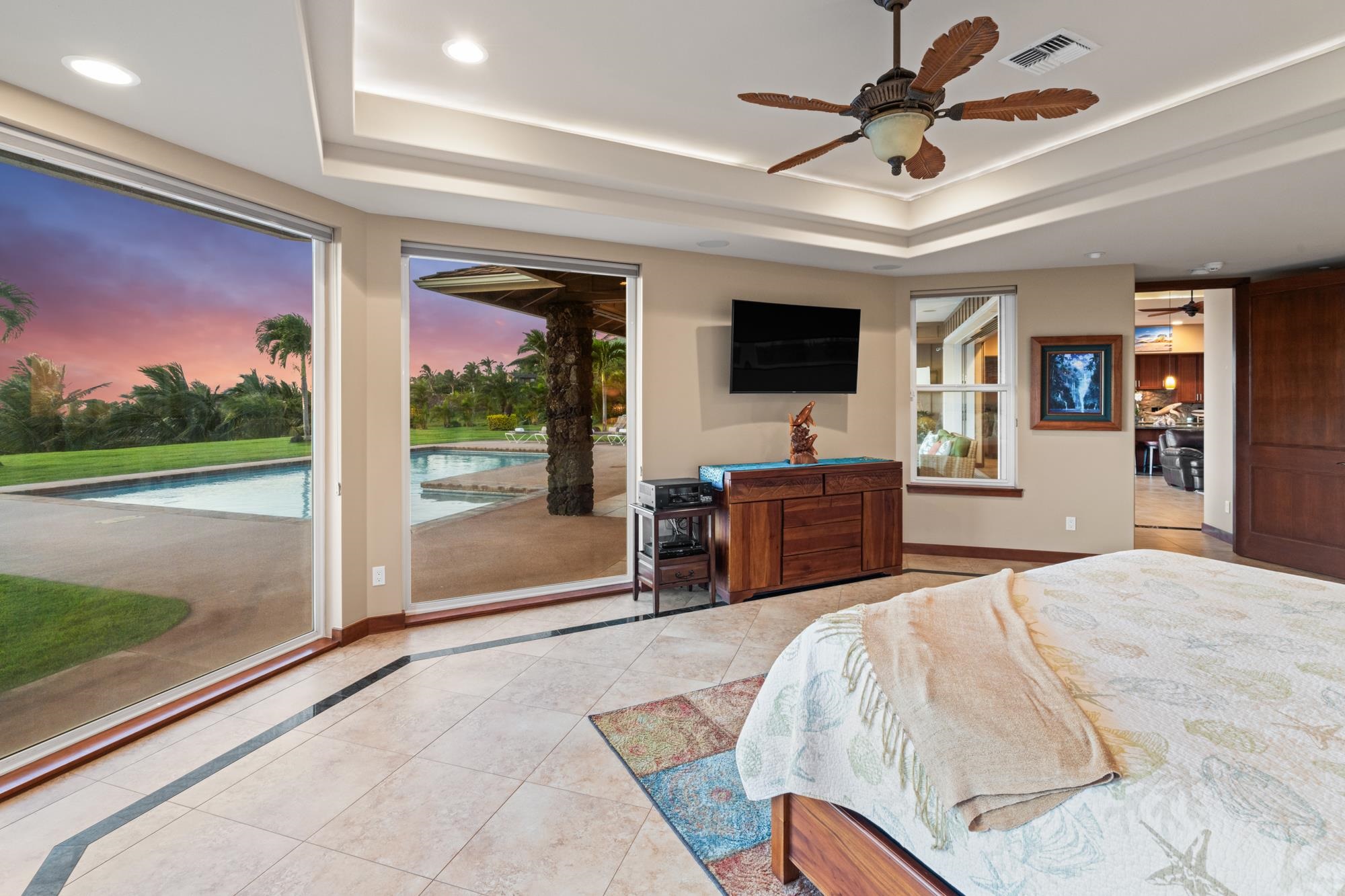 Furnished bedroom with large windows overlooking a pool and tropical landscaping at sunset.