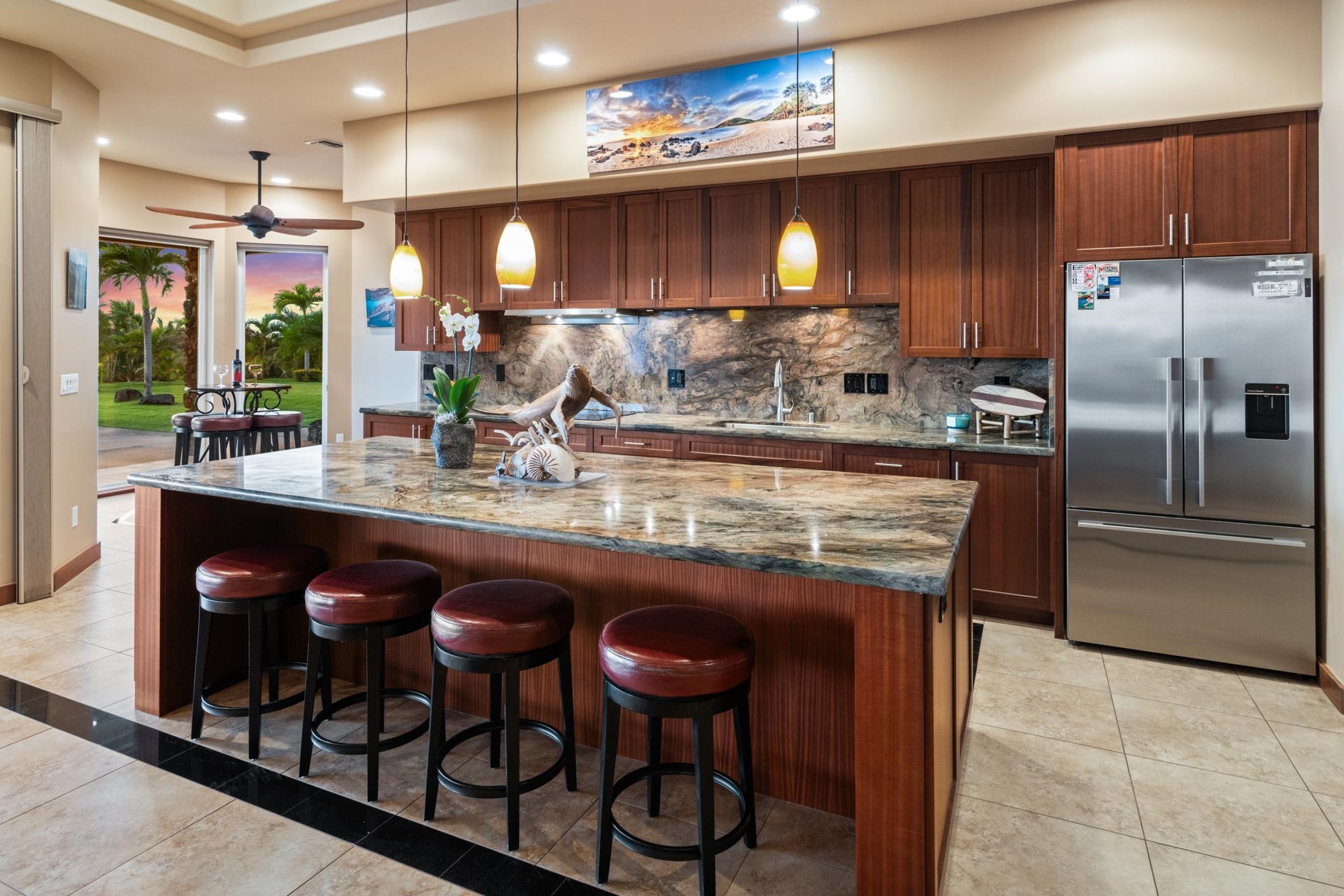 Kitchen with a large stone island, dark wood cabinets, stainless appliances, and sliding doors to a palm-lined lawn.