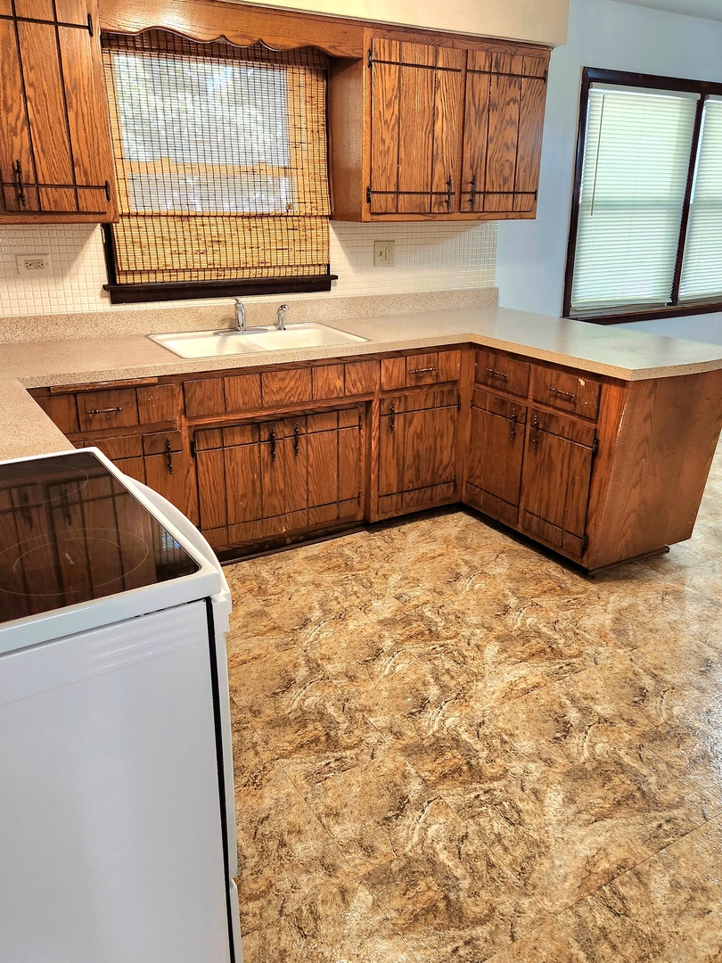 Kitchen with L-shaped wood cabinets, double-basin sink, woven window shade, and patterned flooring.