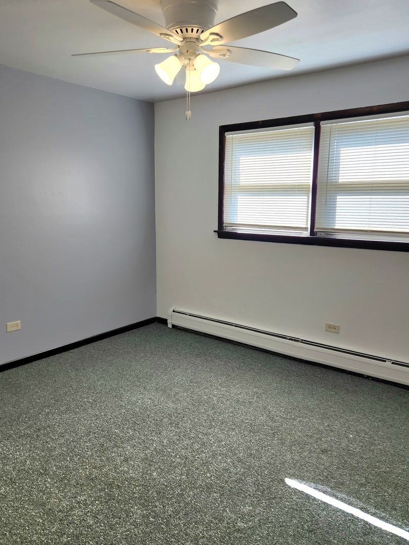 Empty bedroom with gray carpet, a lit ceiling fan, and two windows with blinds.