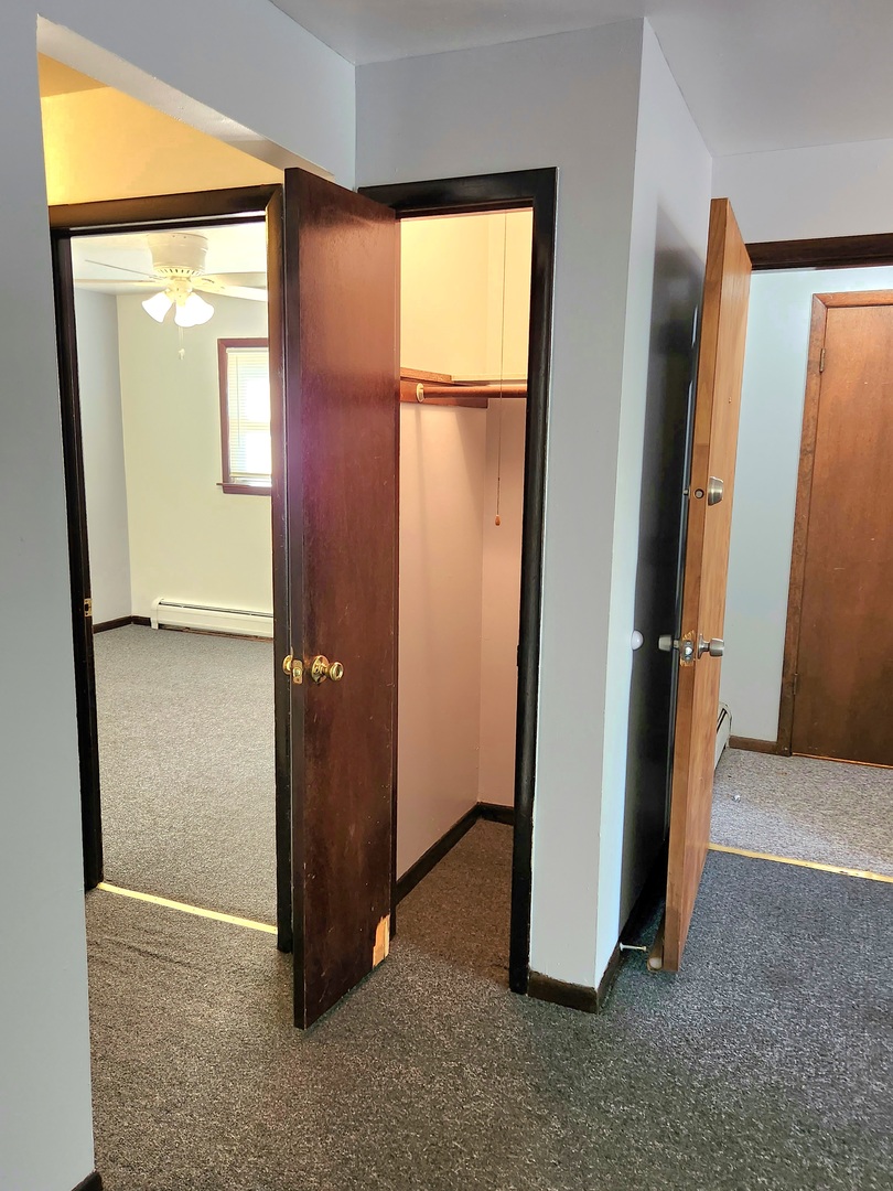 Open closet between carpeted rooms, with dark wood doors and a ceiling fan visible beyond.