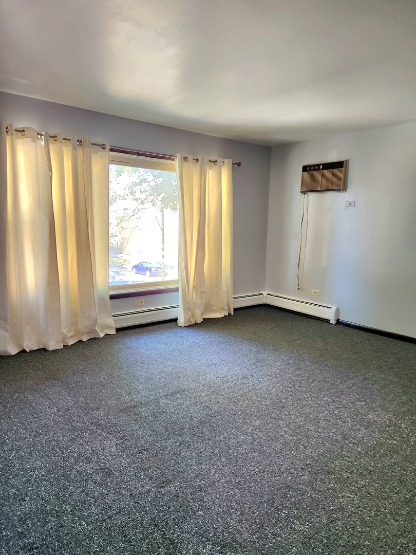 Empty carpeted room with cream curtains, a large window, baseboard heating, and a wall-mounted air conditioner.